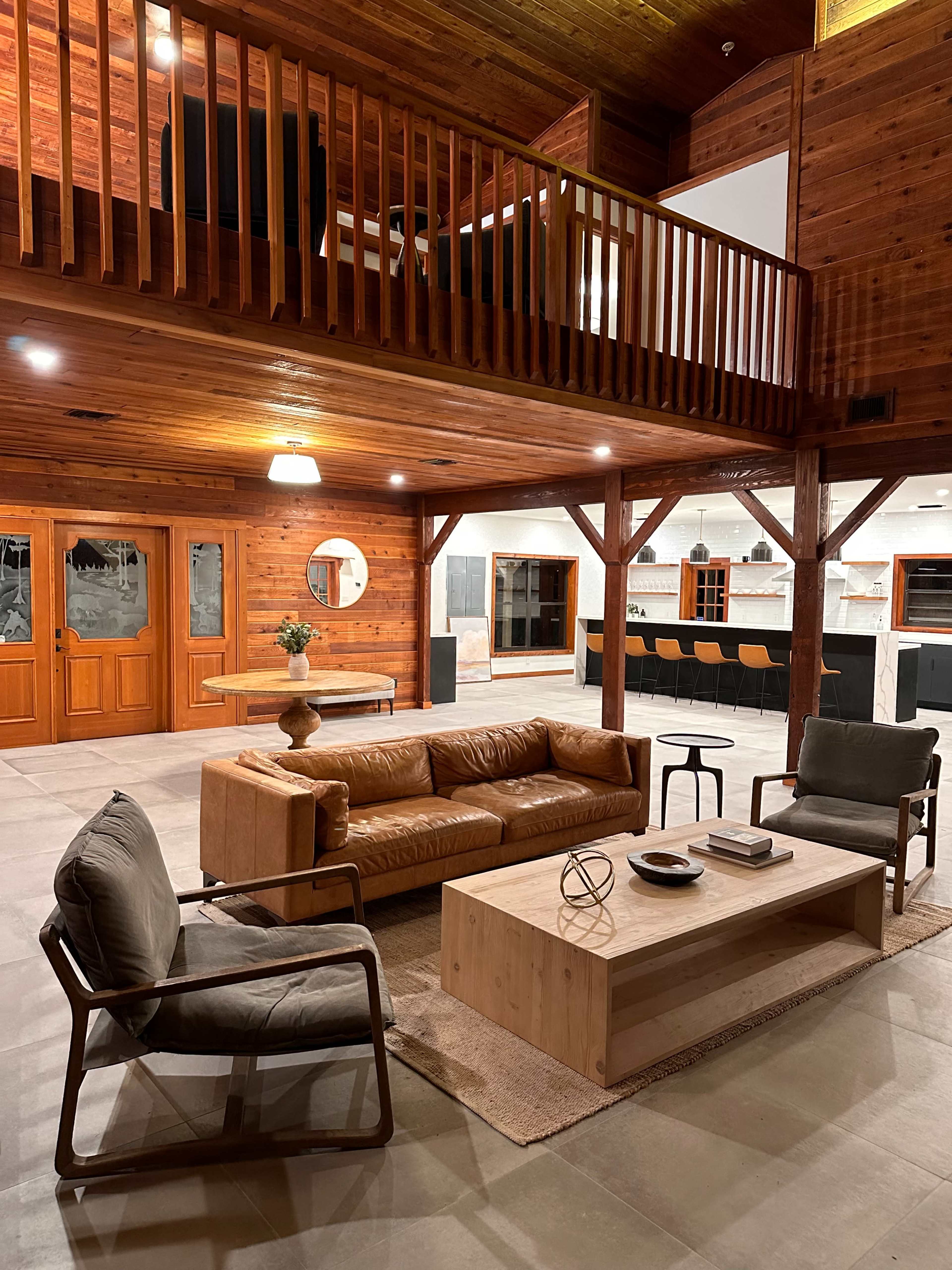 A spacious, modern living area with a wooden loft, featuring a leather sofa, two armchairs, and a coffee table on a light-colored rug.