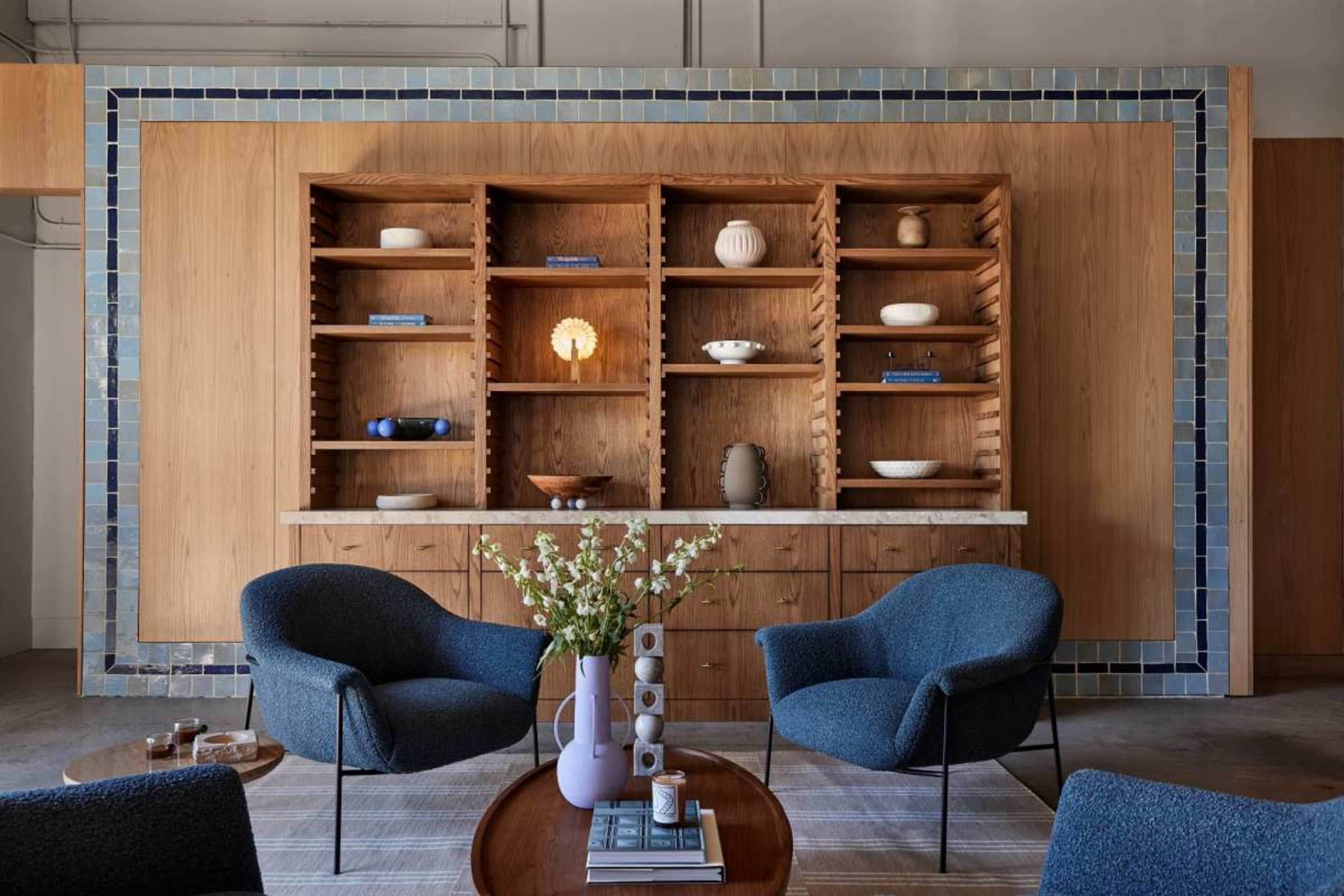 A minimalist interior features a wooden shelving unit filled with decorative items and a low table in front of two blue armchairs.