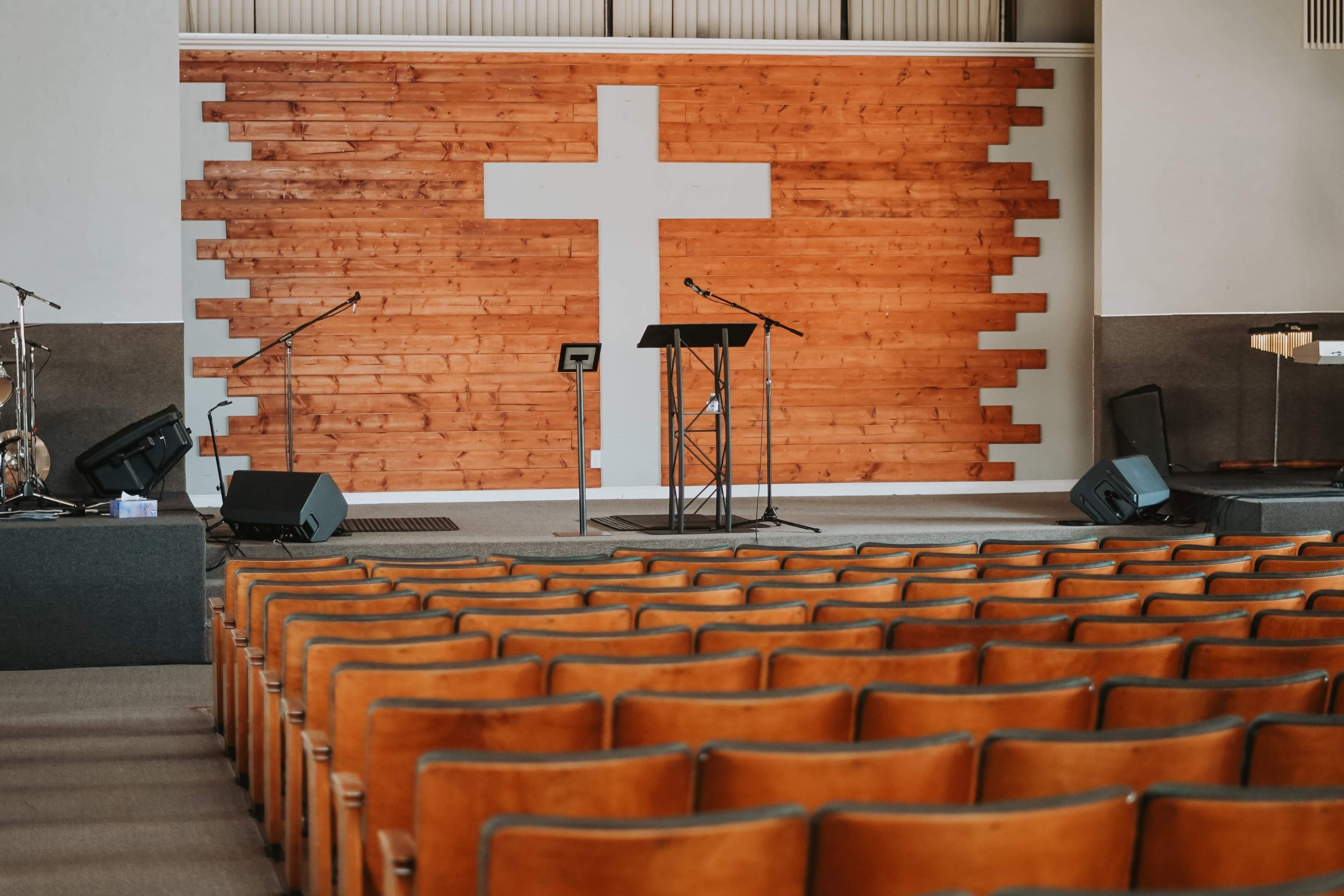 An empty auditorium with rows of orange chairs facing a stage that features a large wooden cross on the wall behind it.