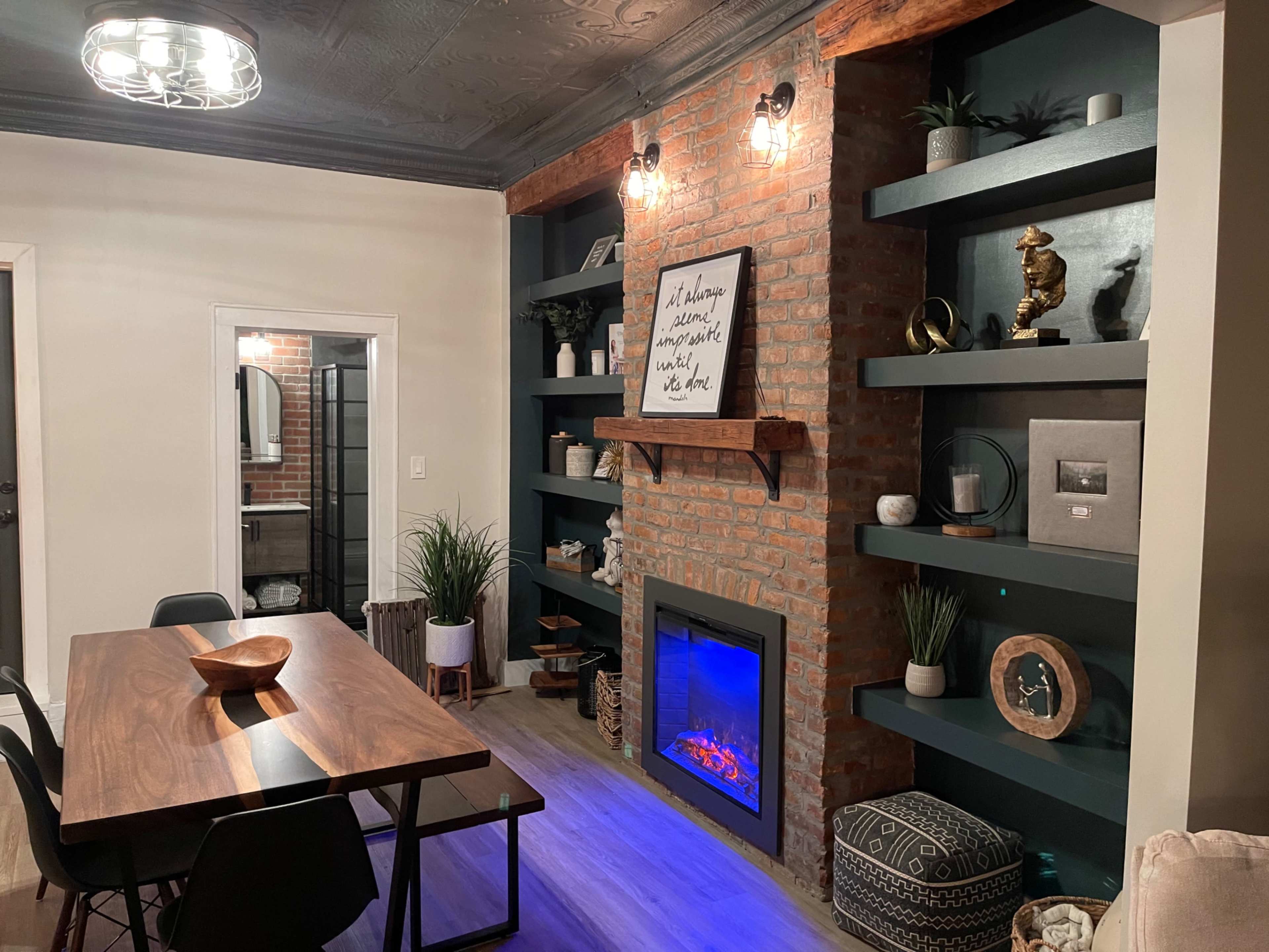 A modern living space featuring a dining table, a wall-mounted fireplace, and built-in shelves with decorative items against a brick wall.