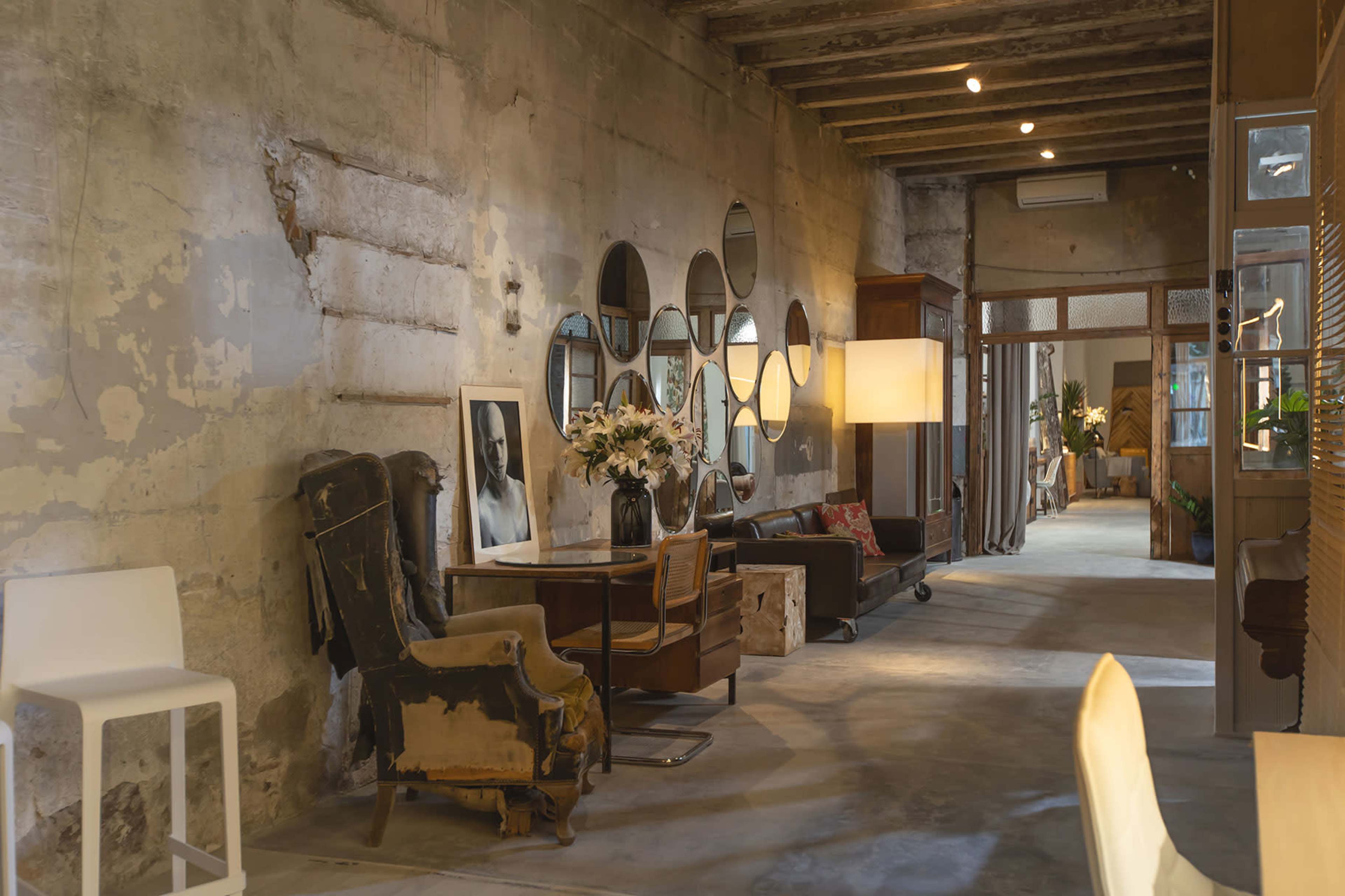 The image shows a modern interior space with exposed concrete walls, decorative mirrors, and a mix of vintage and contemporary furniture.