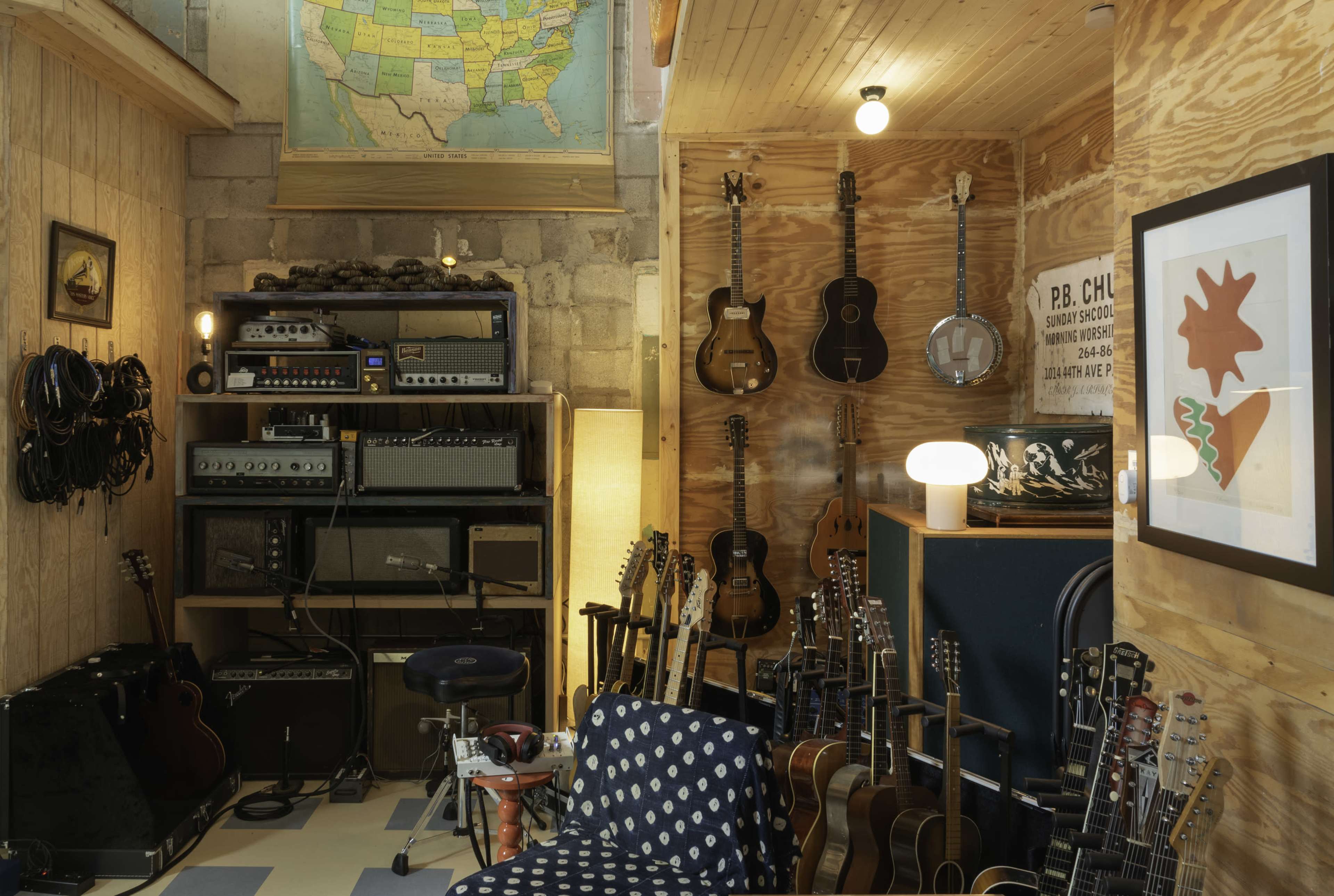 The image shows a room filled with musical instruments, including guitars neatly arranged on the walls and amplifiers set up against a wooden backdrop.