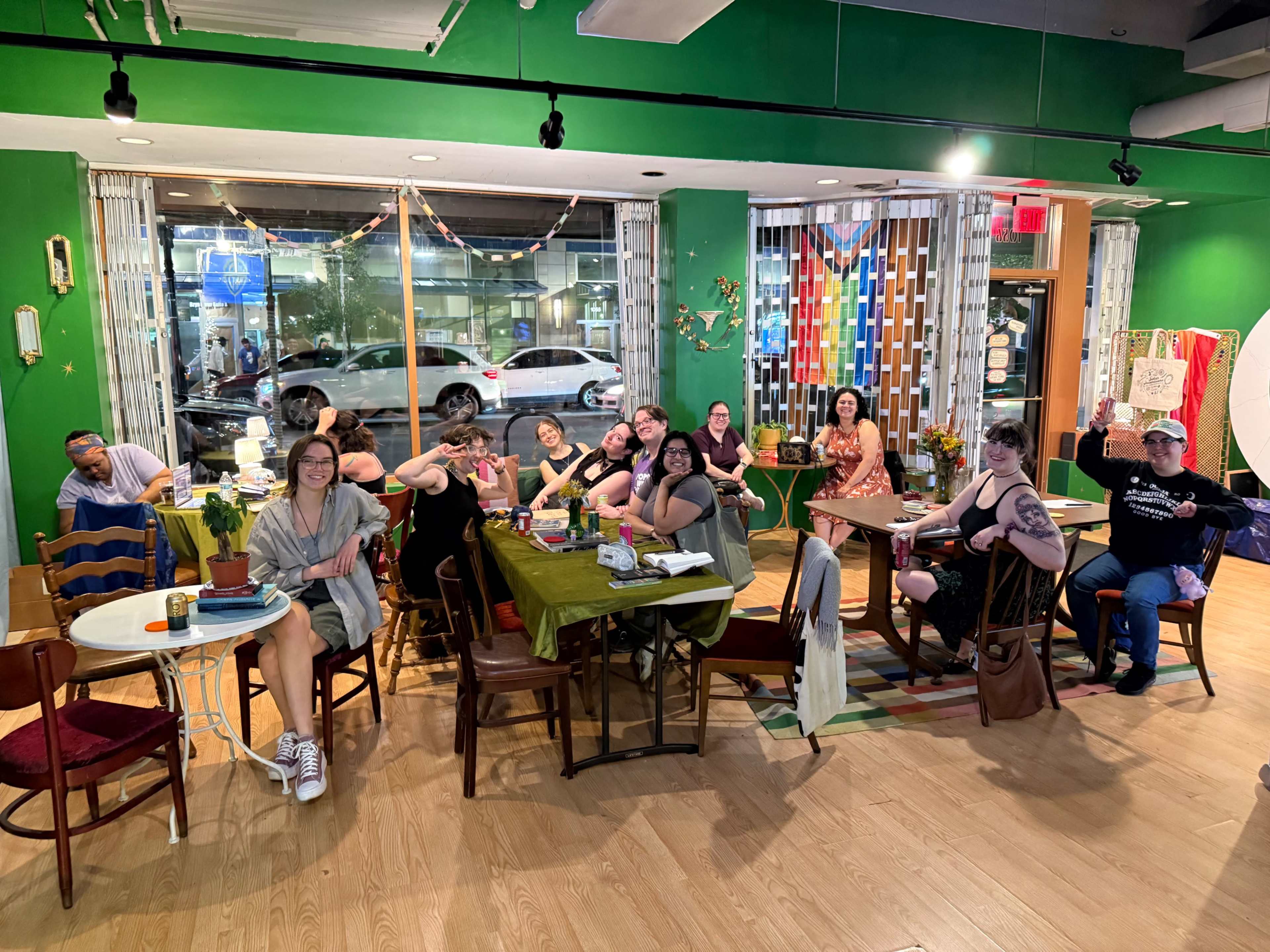 A diverse group of people gathers at various tables in a brightly colored café, with plants and decorations visible in the background.