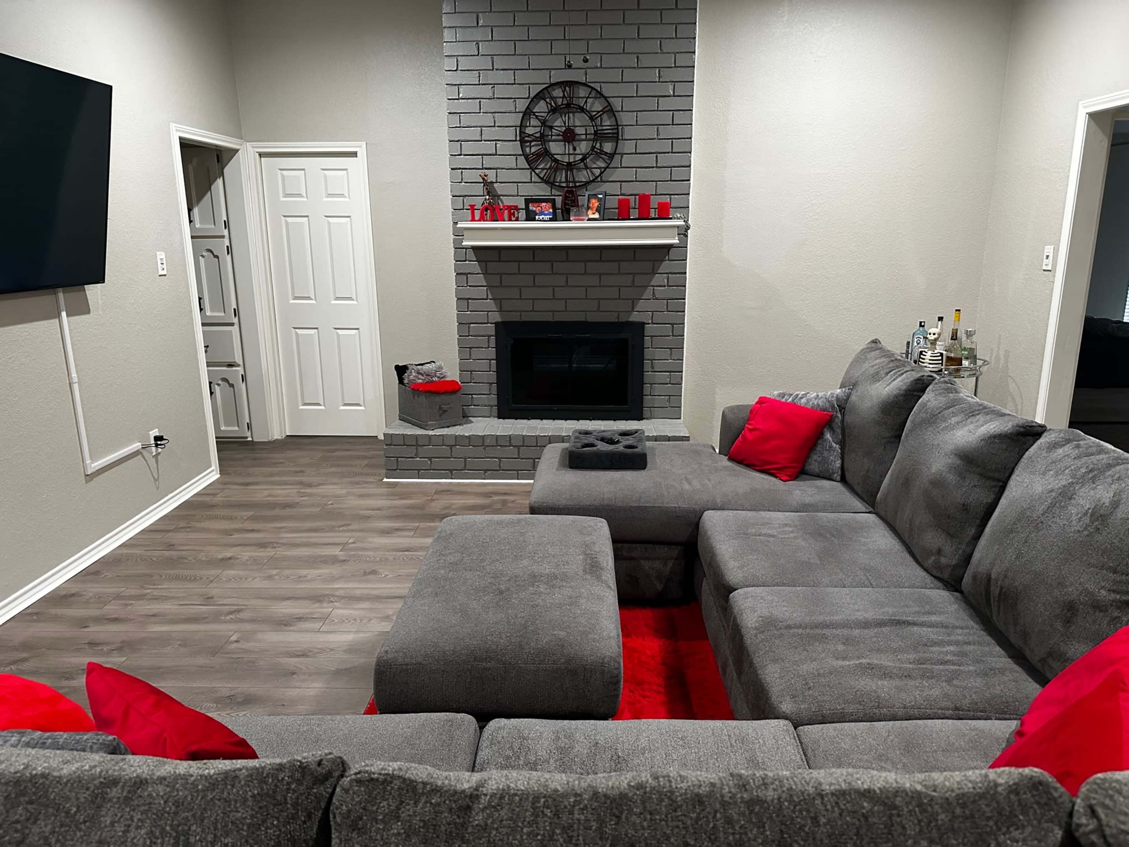 A living room with a gray sectional sofa, a dark gray fireplace with a decorative clock above it, and a television mounted on the wall.