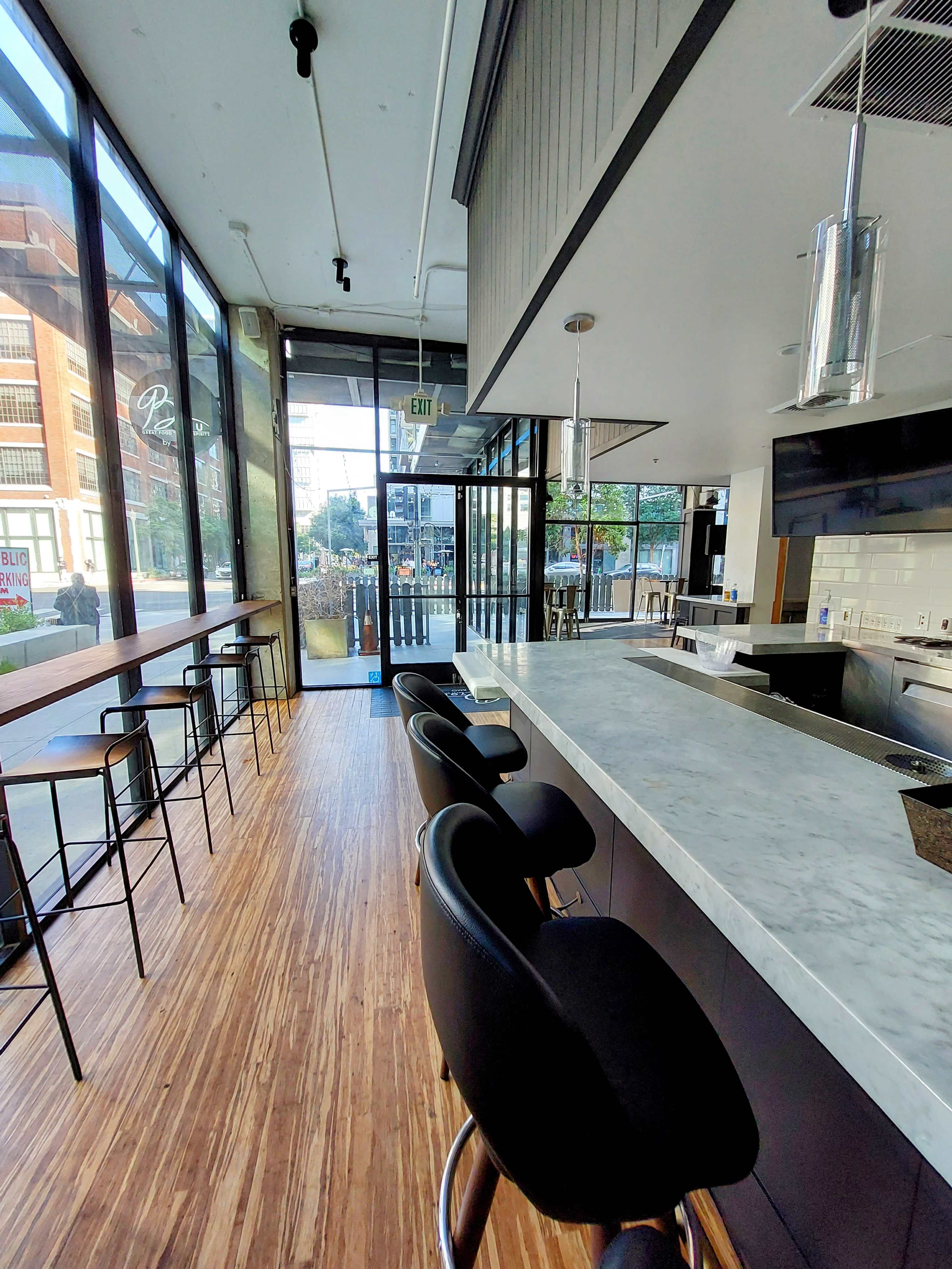 A modern restaurant interior featuring a marble-topped bar with black stools and large windows that provide a view of the outdoor seating area.