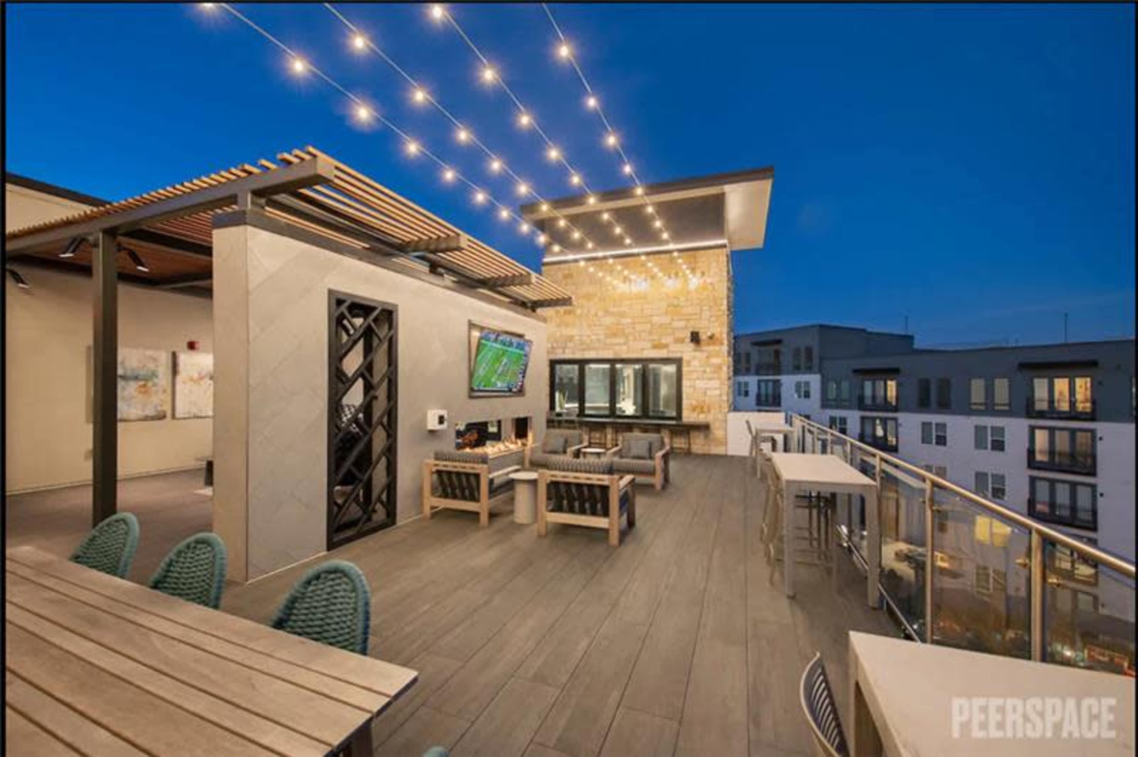 The image shows a rooftop terrace with string lights, outdoor seating, and a wall-mounted TV, overlooking a modern building at dusk.