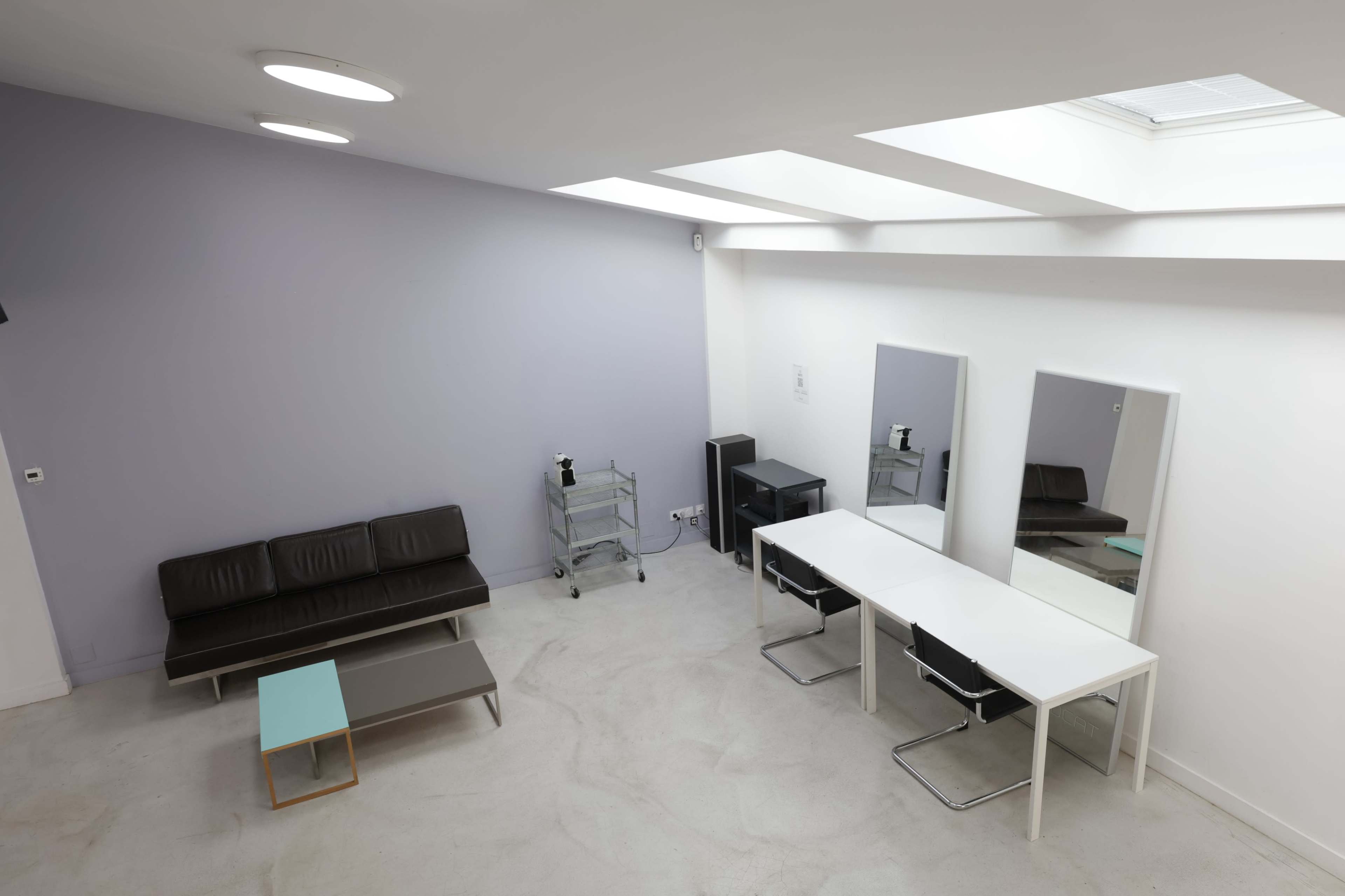 The interior space features a minimalist design with a brown sofa, a glass-top coffee table, a white table with black chairs, and two mirrors against a pale wall, illuminated by natural light from skylights.