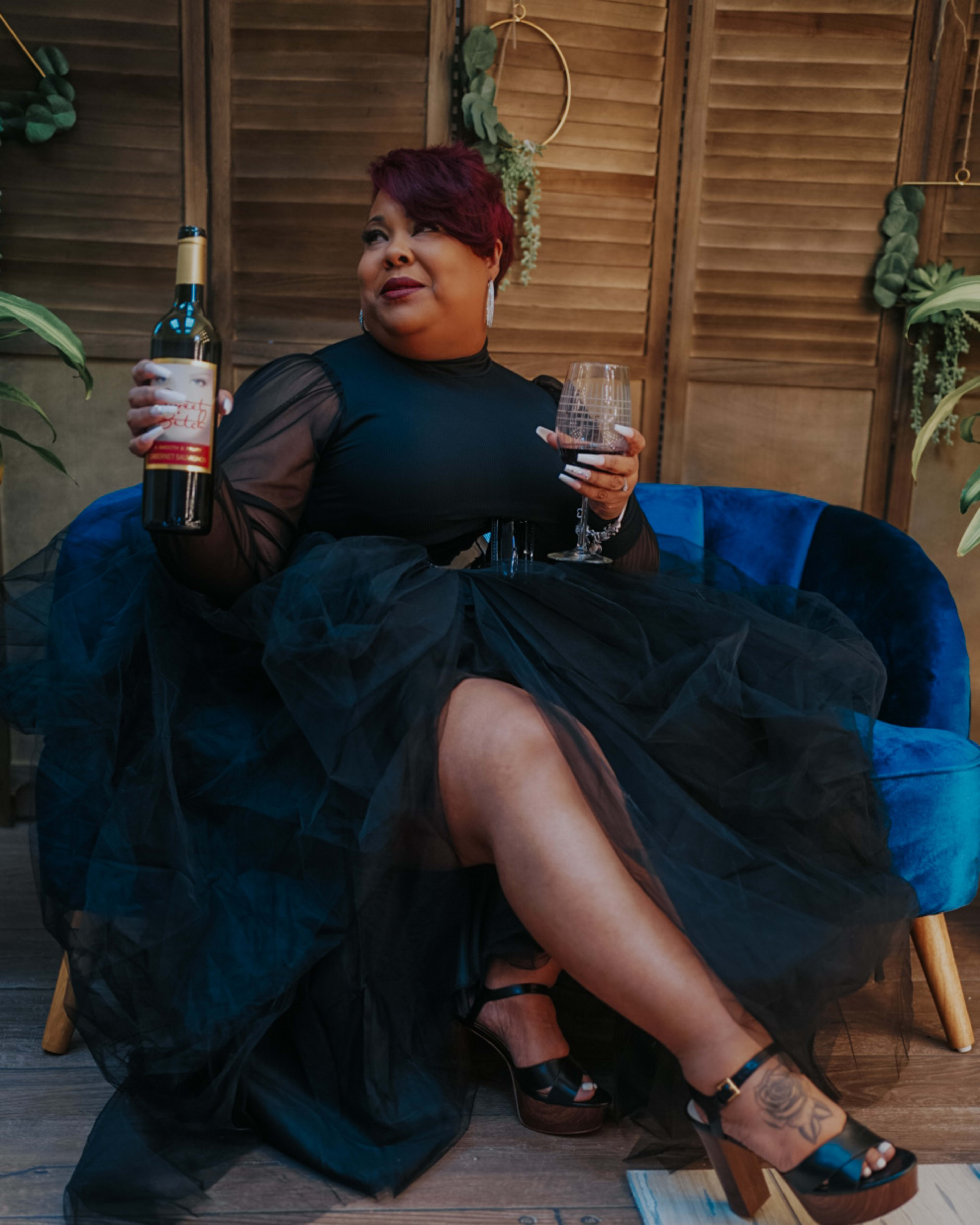 Una mujer sosteniendo una botella de vino sentada en una silla azul durante una sesión de fotos.