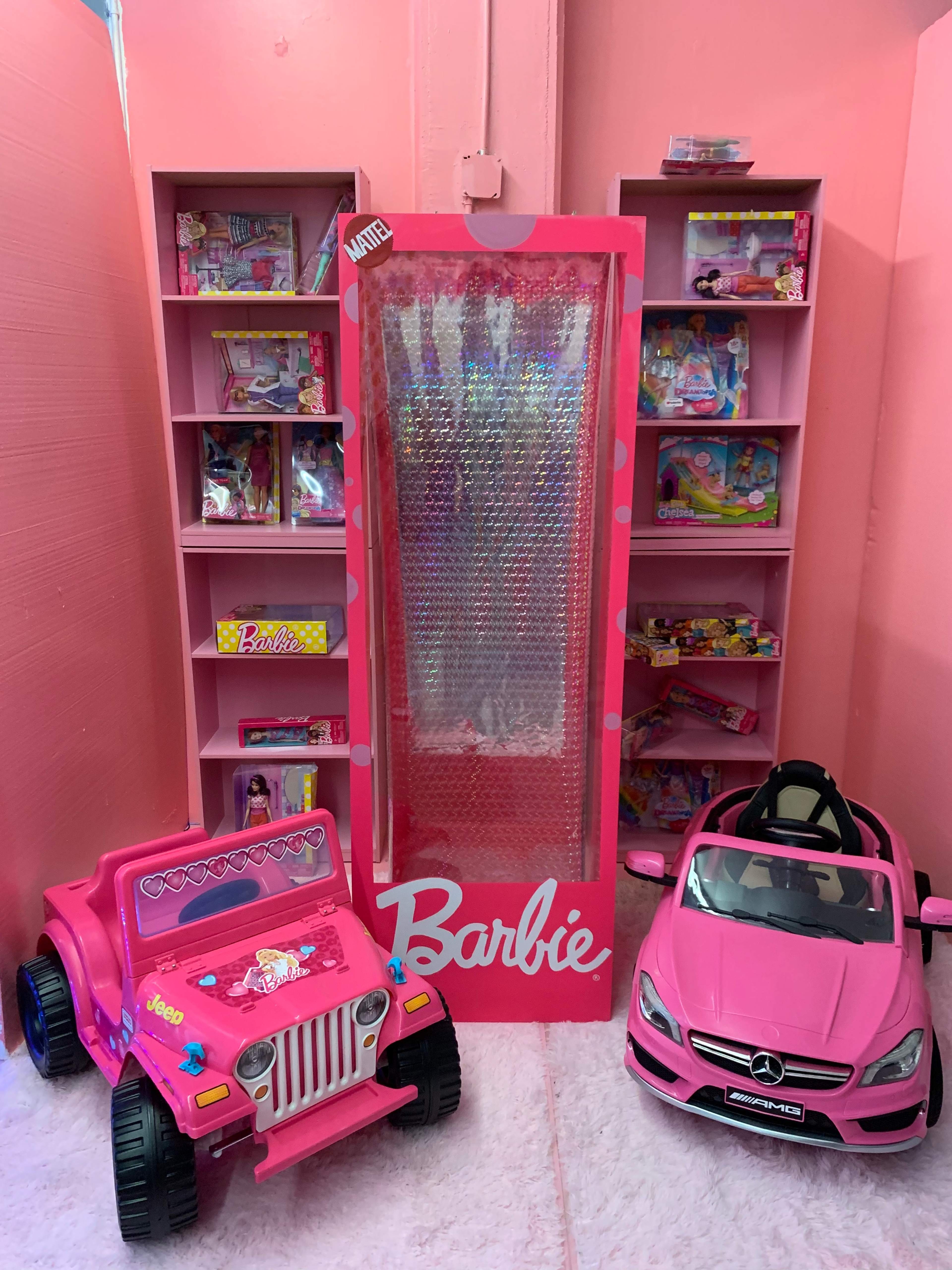 The image features a pink-themed display with a glass-fronted cabinet filled with Barbie toys, alongside two pink toy cars, a jeep and a luxury sports car.