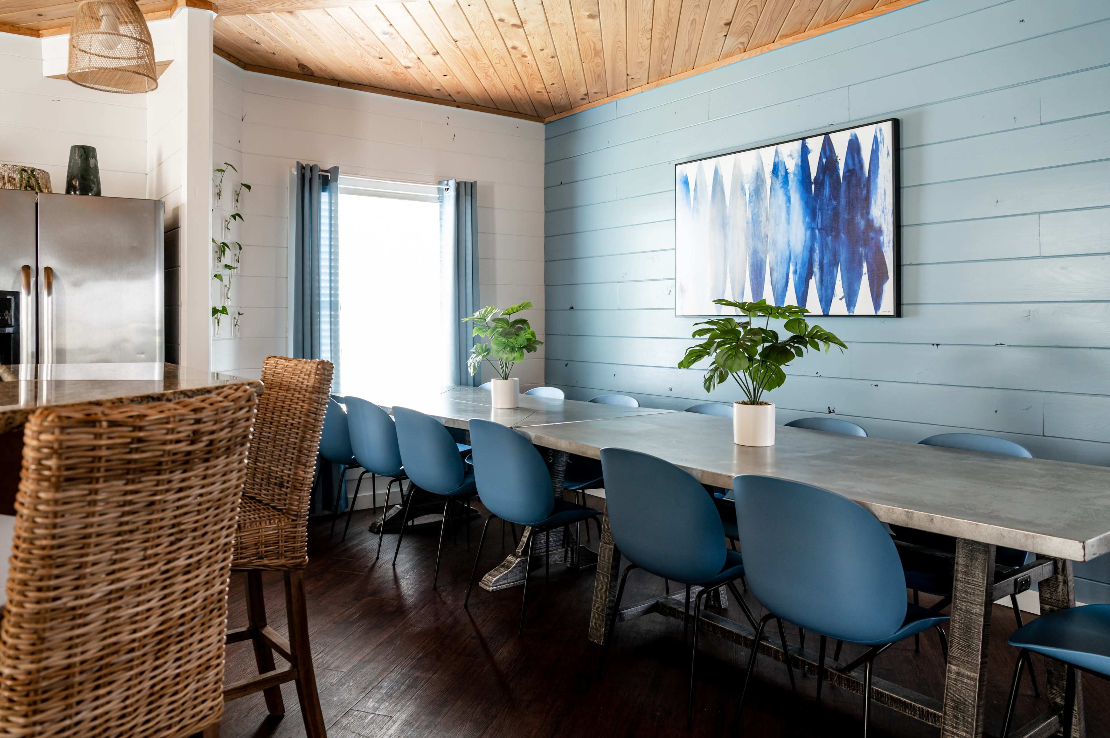 A long table with blue chairs is set in a dining area featuring light blue walls and a large window with curtains.