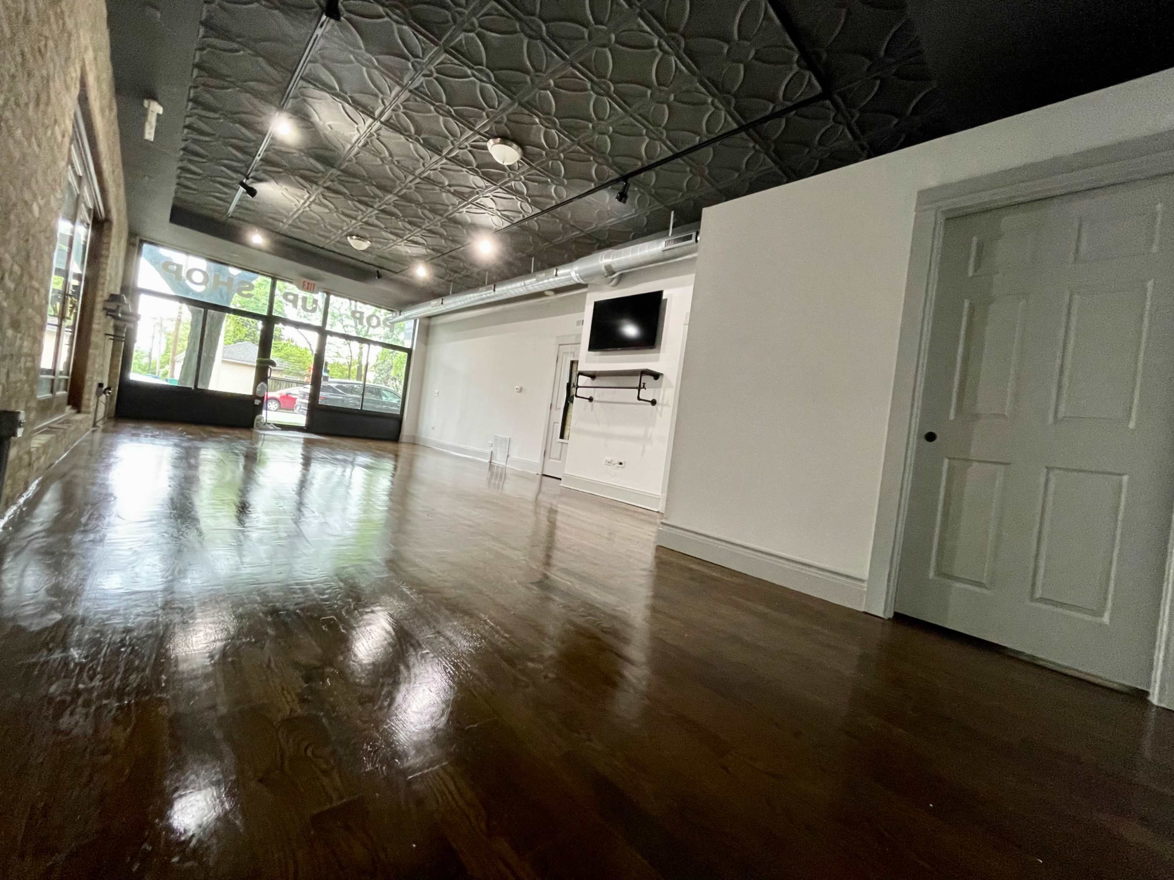 An empty room with polished wooden floors, a ceiling with decorative tiles, and large windows facing the street.