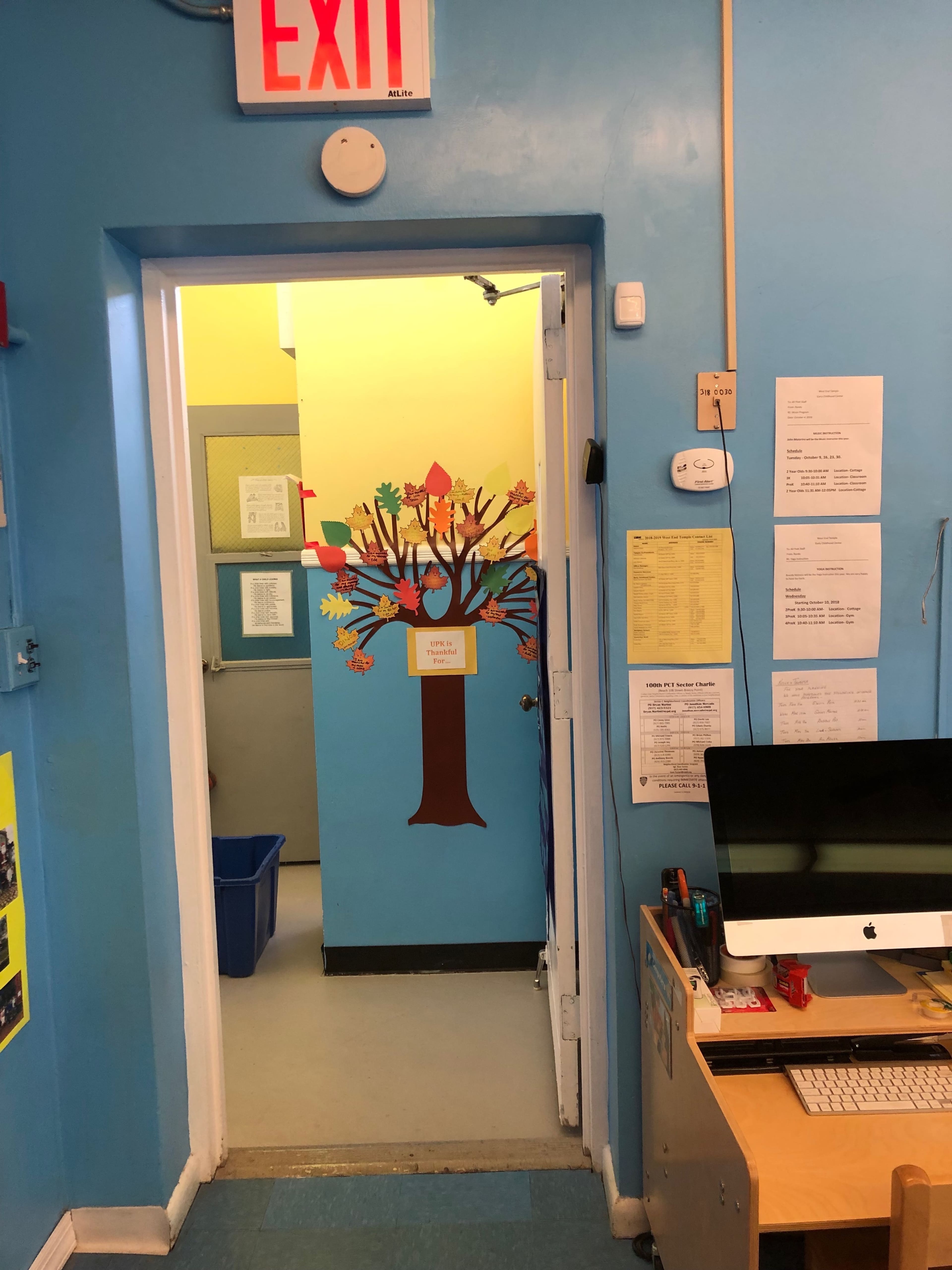 The image shows a brightly colored classroom interior with a door leading to another room, featuring a tree mural on the wall and an exit sign above.