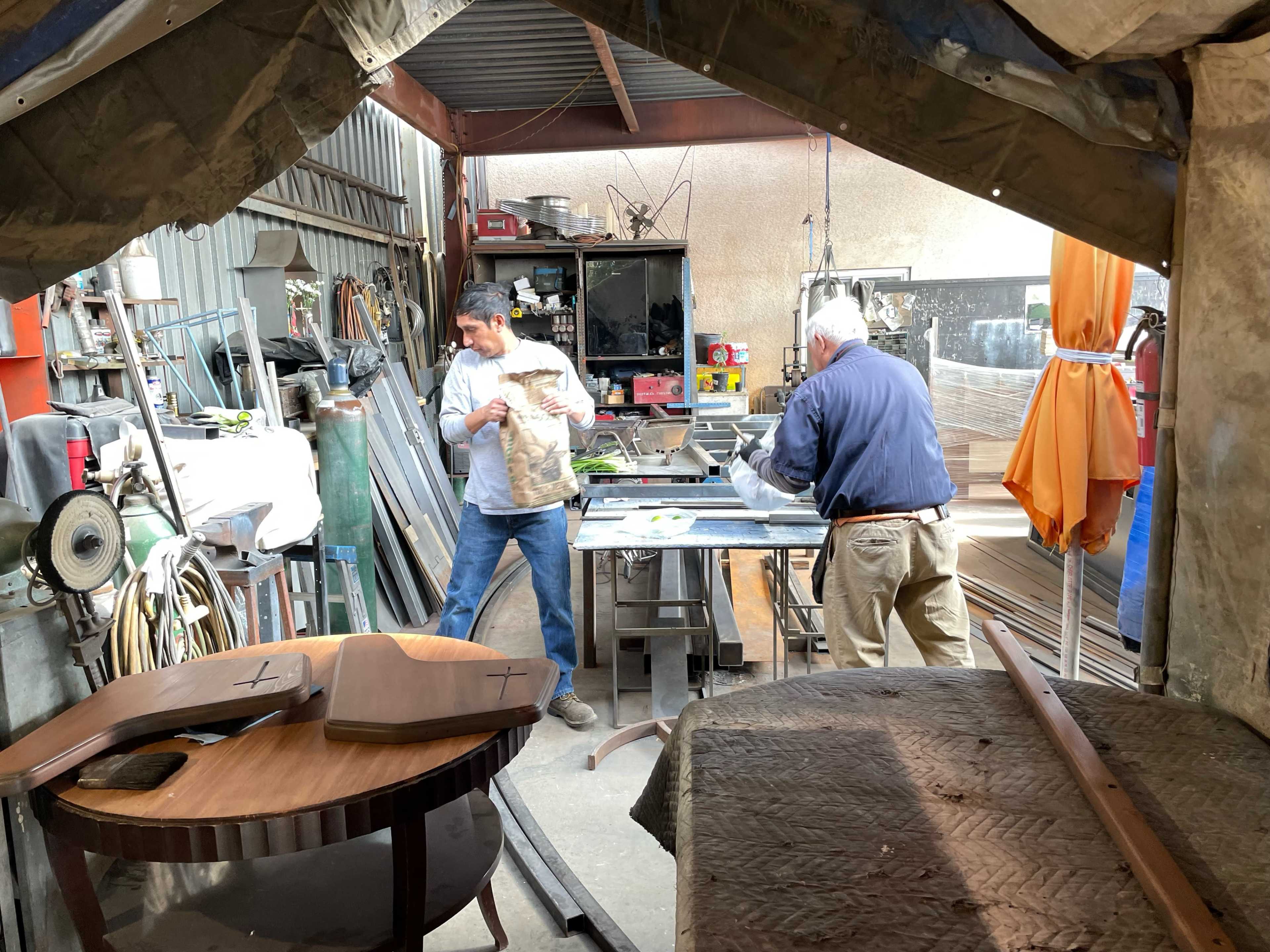 Two men are working in a workshop filled with tools and materials for woodcraft, with one man handling a bag and the other focused on a workbench.