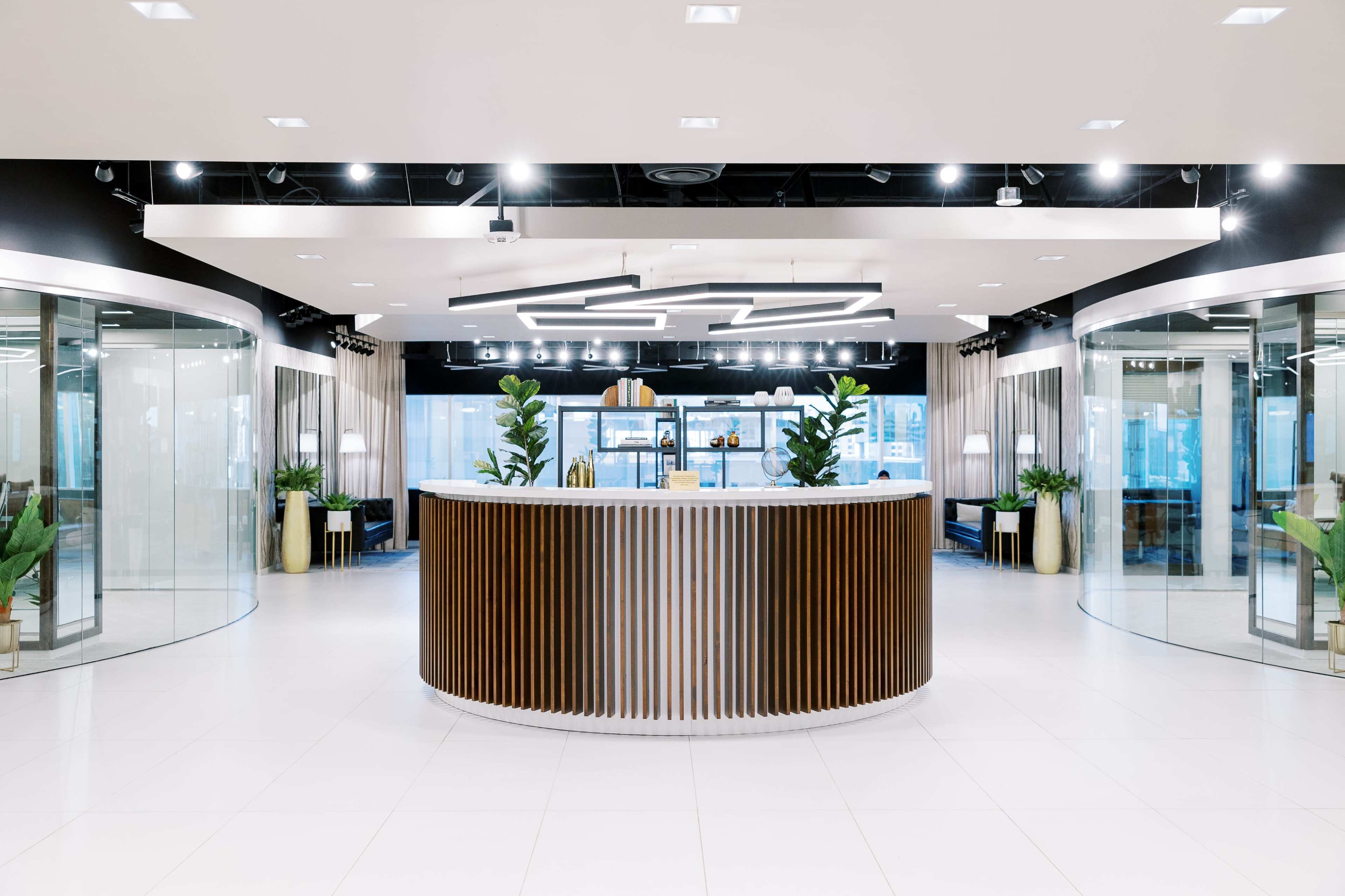 The image shows a modern reception area with a round wooden front desk surrounded by glass walls and decorative plants.