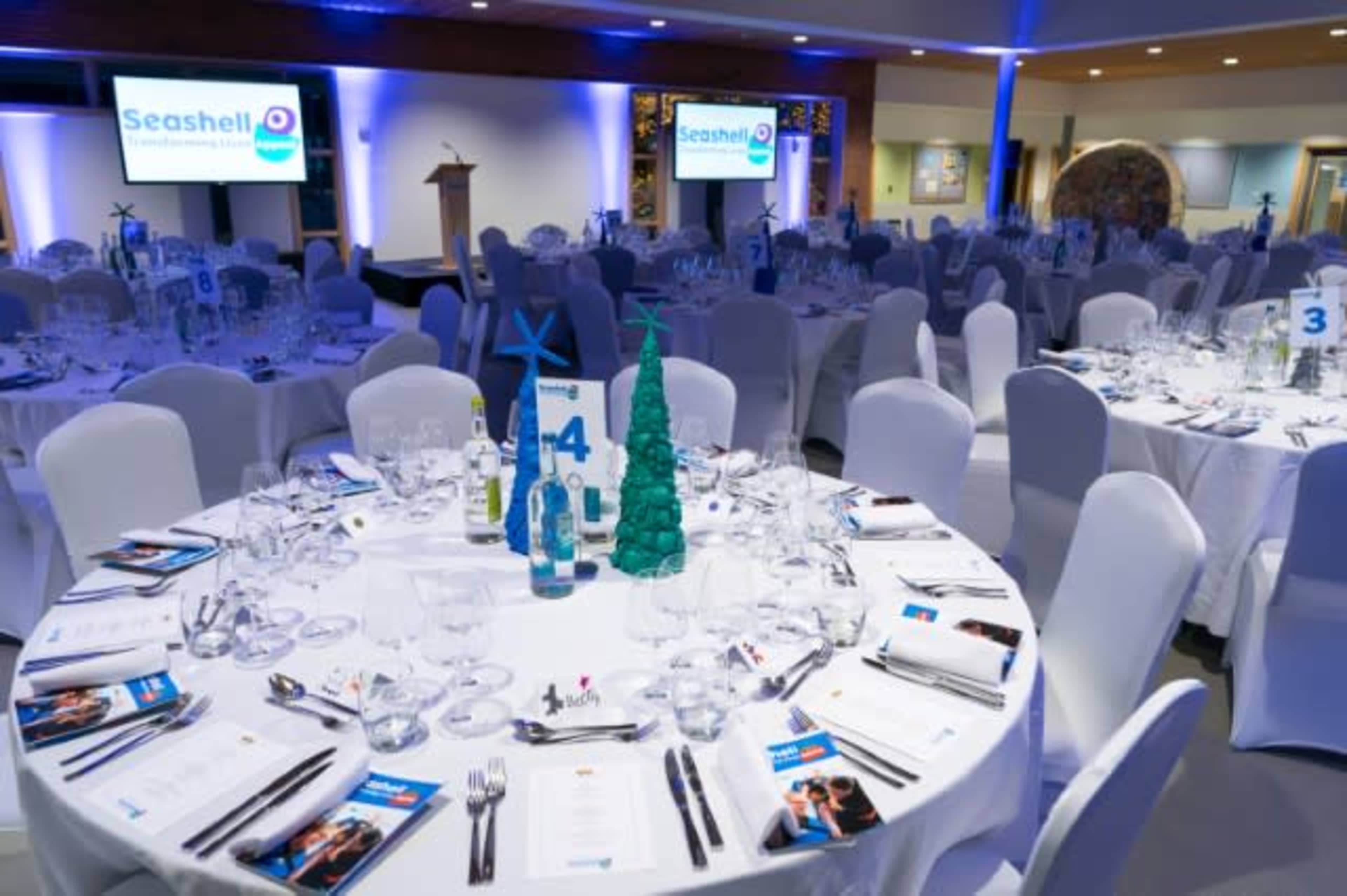 A banquet room is set up for an event with elegantly arranged tables, each featuring place settings and decorative centerpieces.