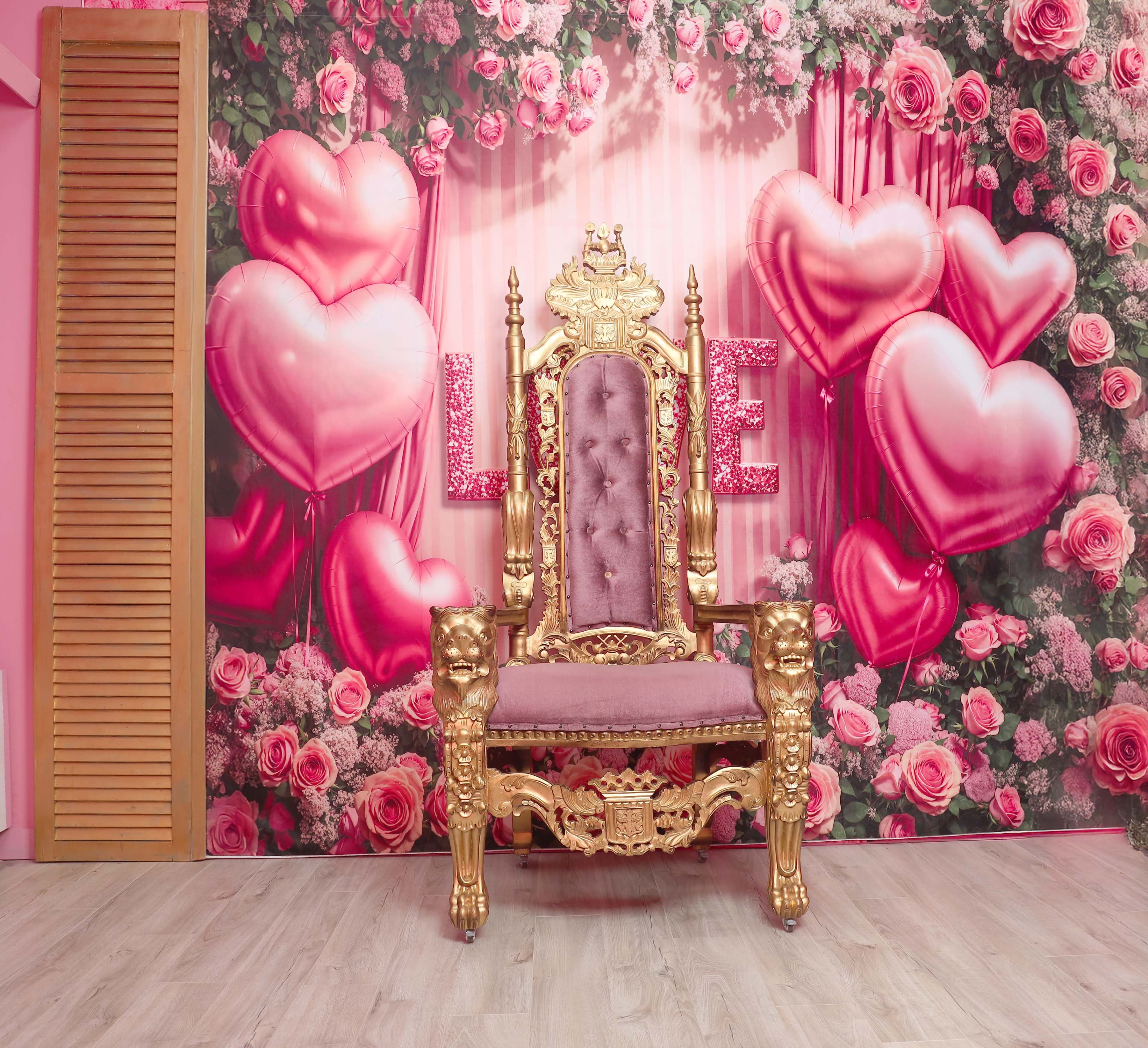 The image features an ornate gold throne with purple upholstery against a pink floral backdrop decorated with heart-shaped balloons.