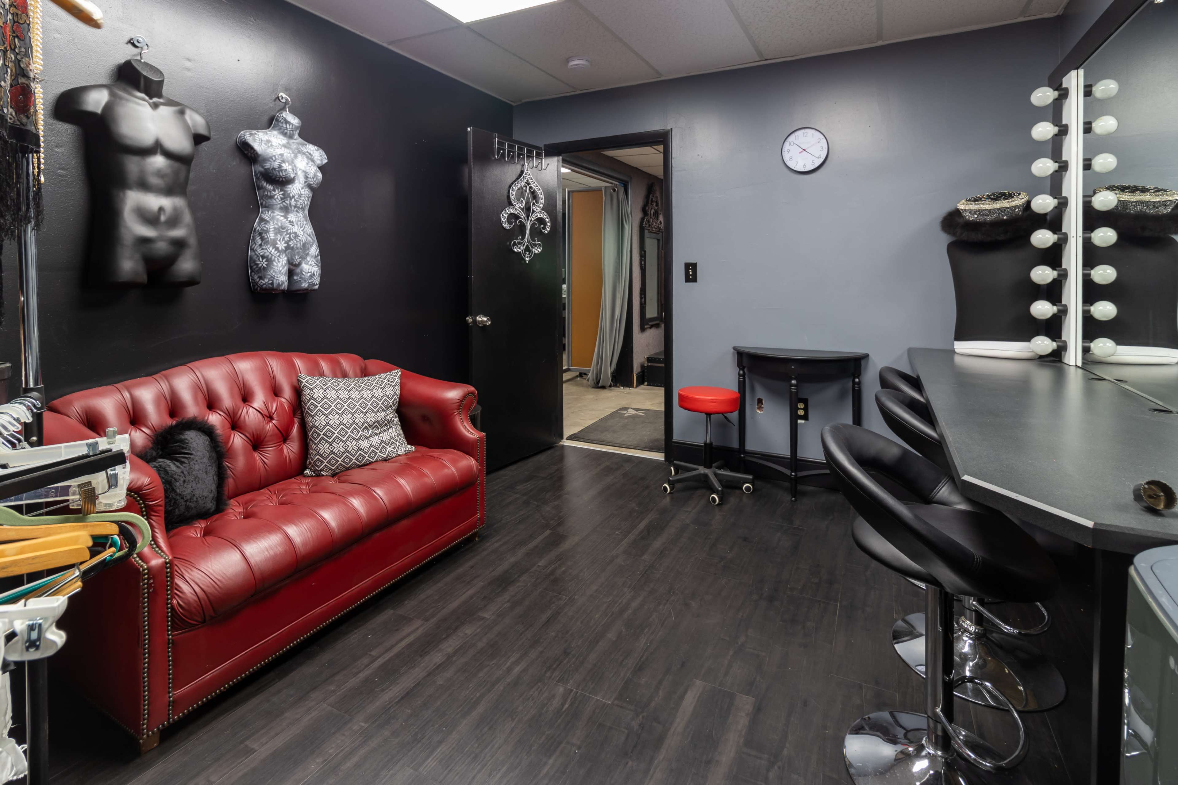 A room with black walls featuring a red leather couch, a small black table, three black barstools, and a mirror with light bulbs, along with mannequins on display.