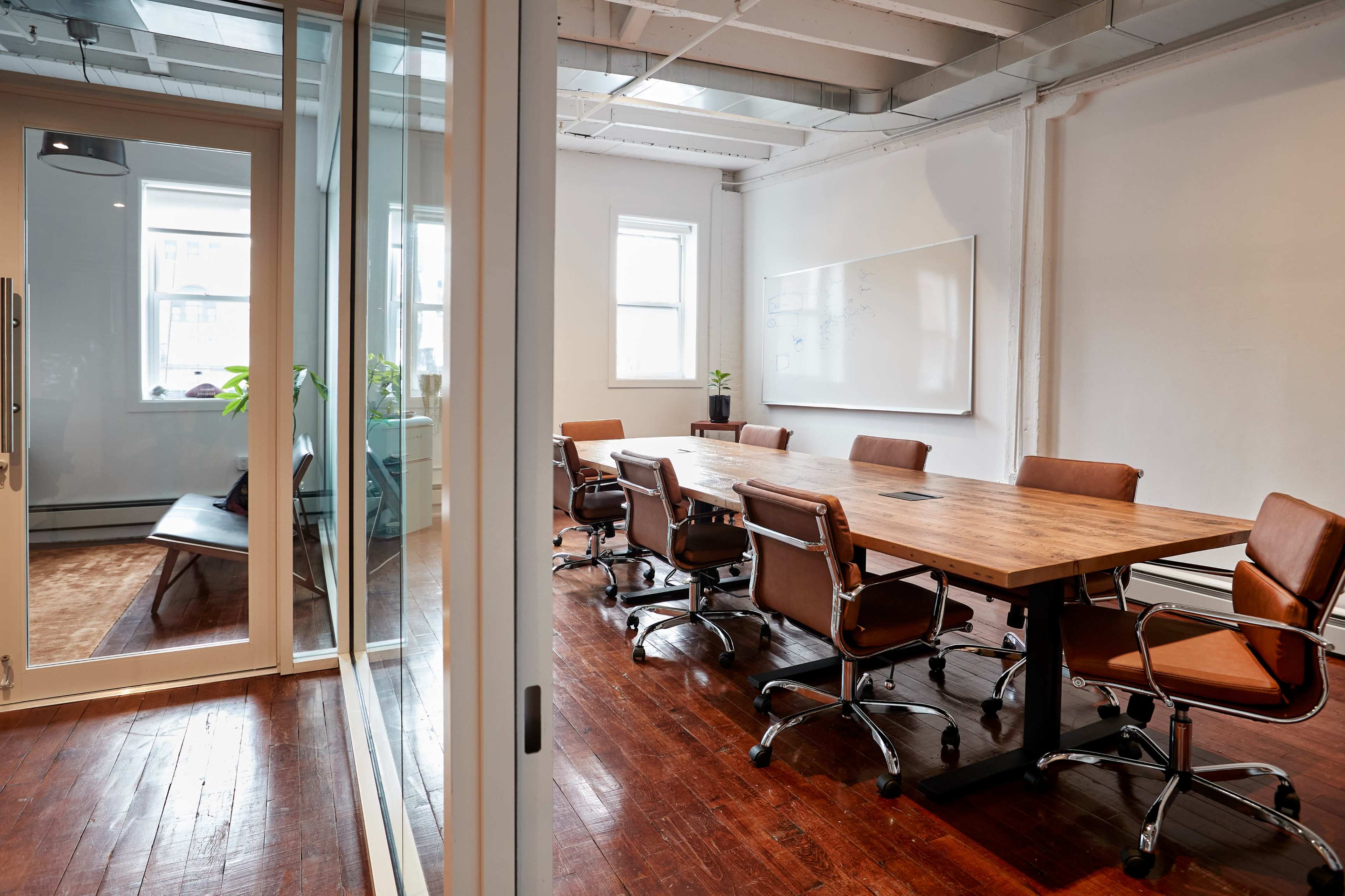 A spacious conference room with a long wooden table surrounded by brown leather swivel chairs, leading to a glass-walled meeting area.