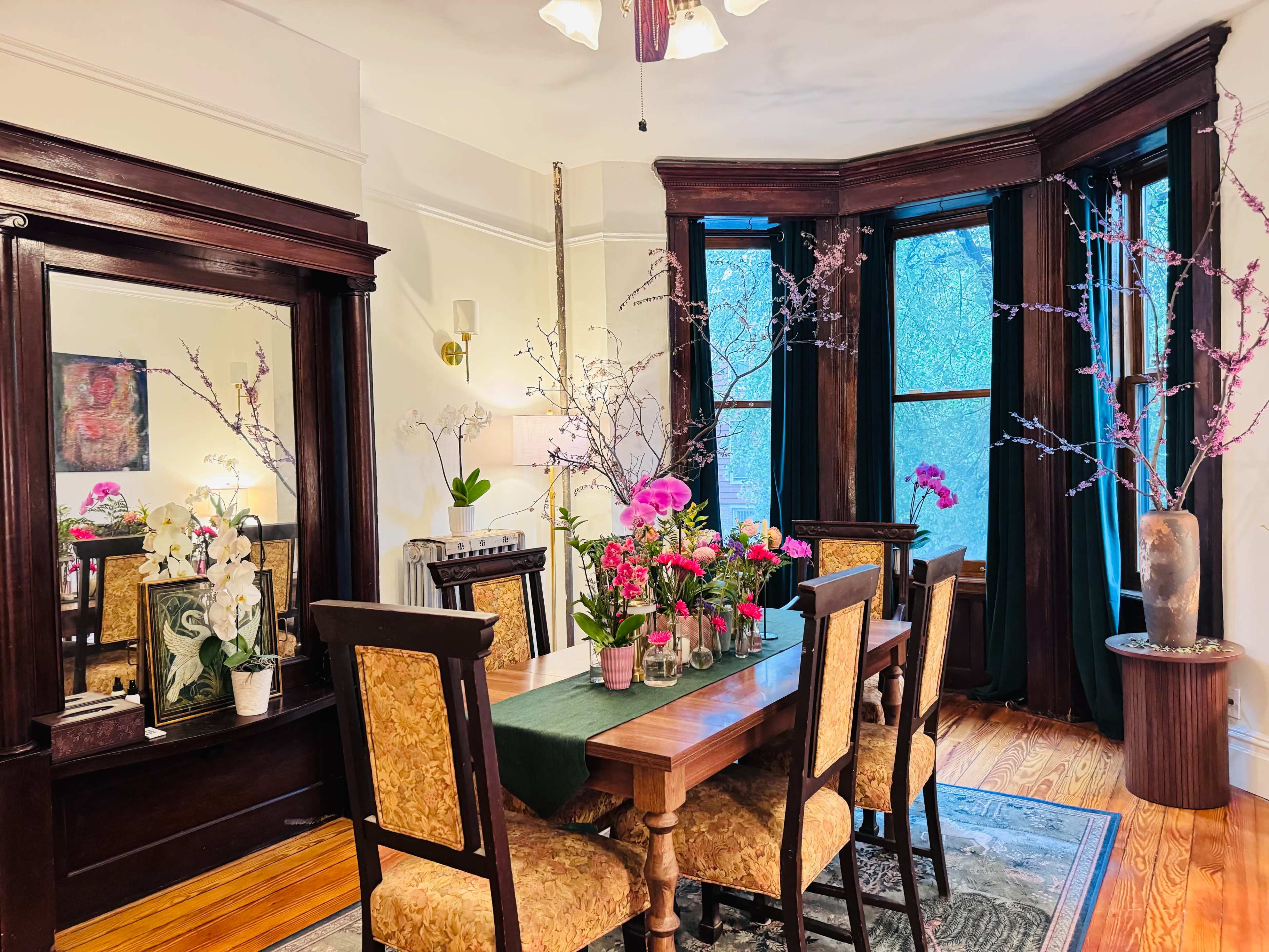 A wooden dining table with chairs is set in a well-lit room featuring large windows, a mirror, and decorative flowers in vases.