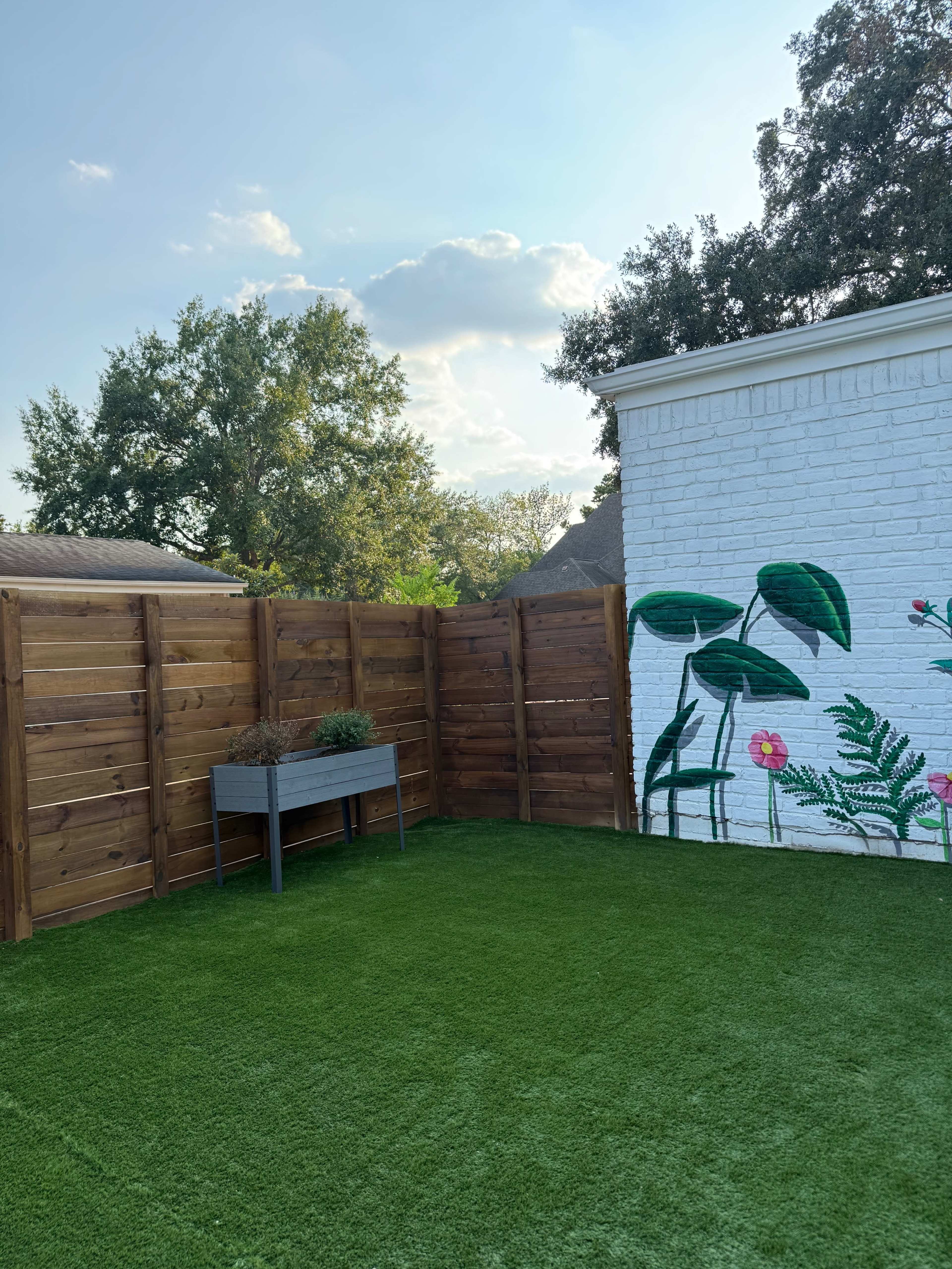 The image shows a backyard with artificial grass, a wooden fence, and a white wall featuring a colorful mural of plants and flowers.