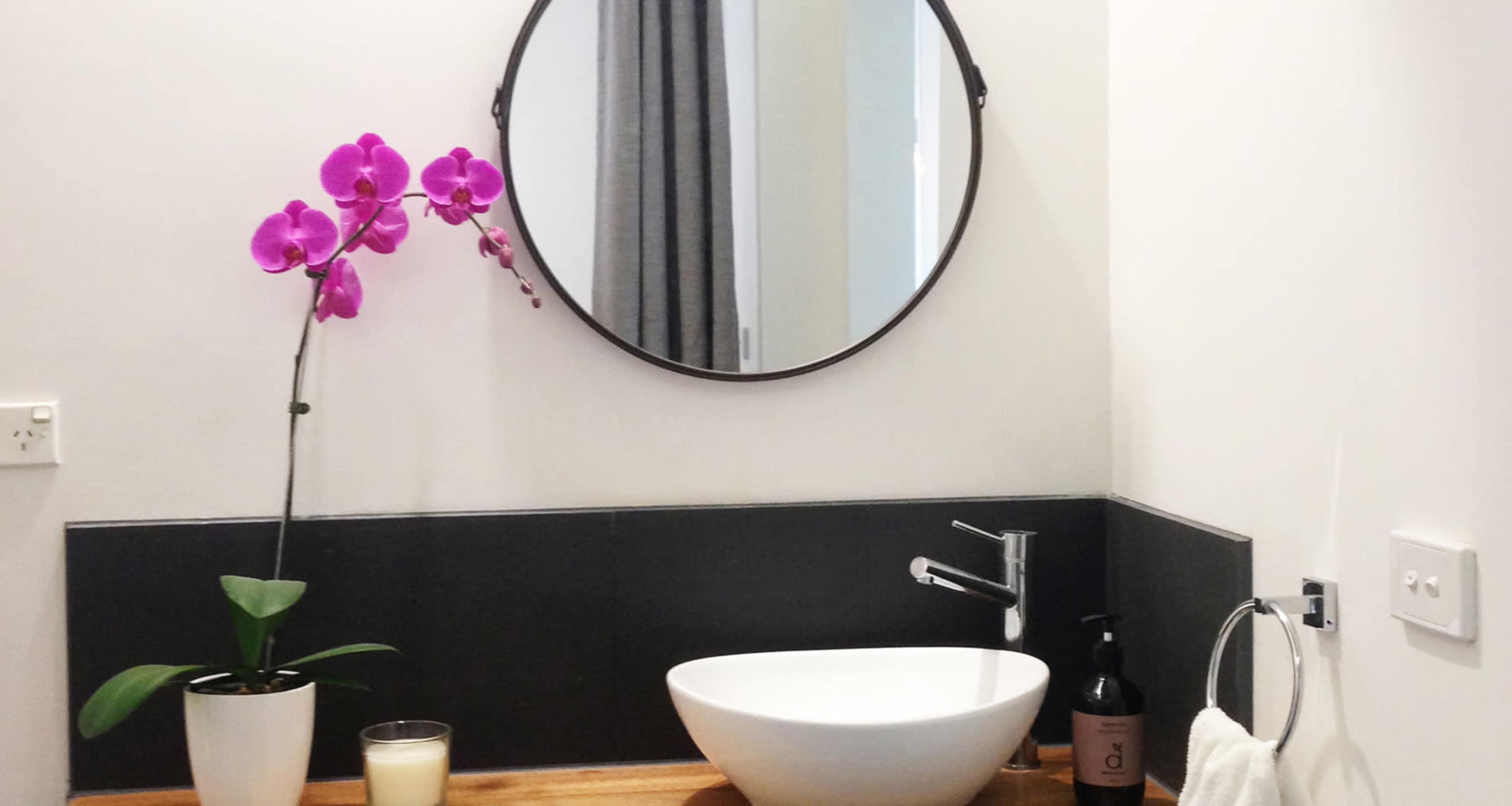 A modern bathroom with a round mirror, a sink, a potted orchid, and a candle on a wooden countertop.