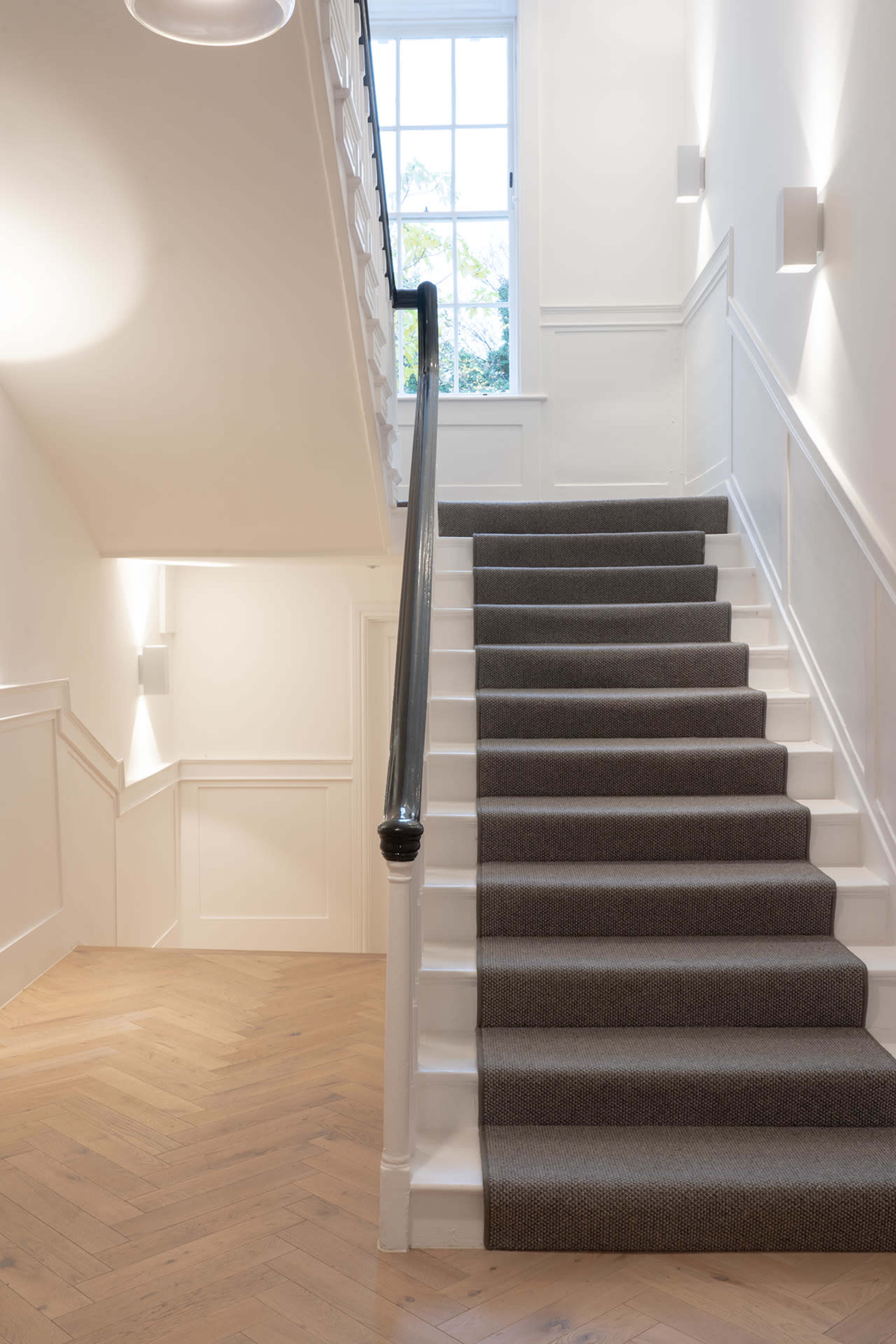 The image shows a well-lit interior staircase with a carpeted path leading up to a window.
