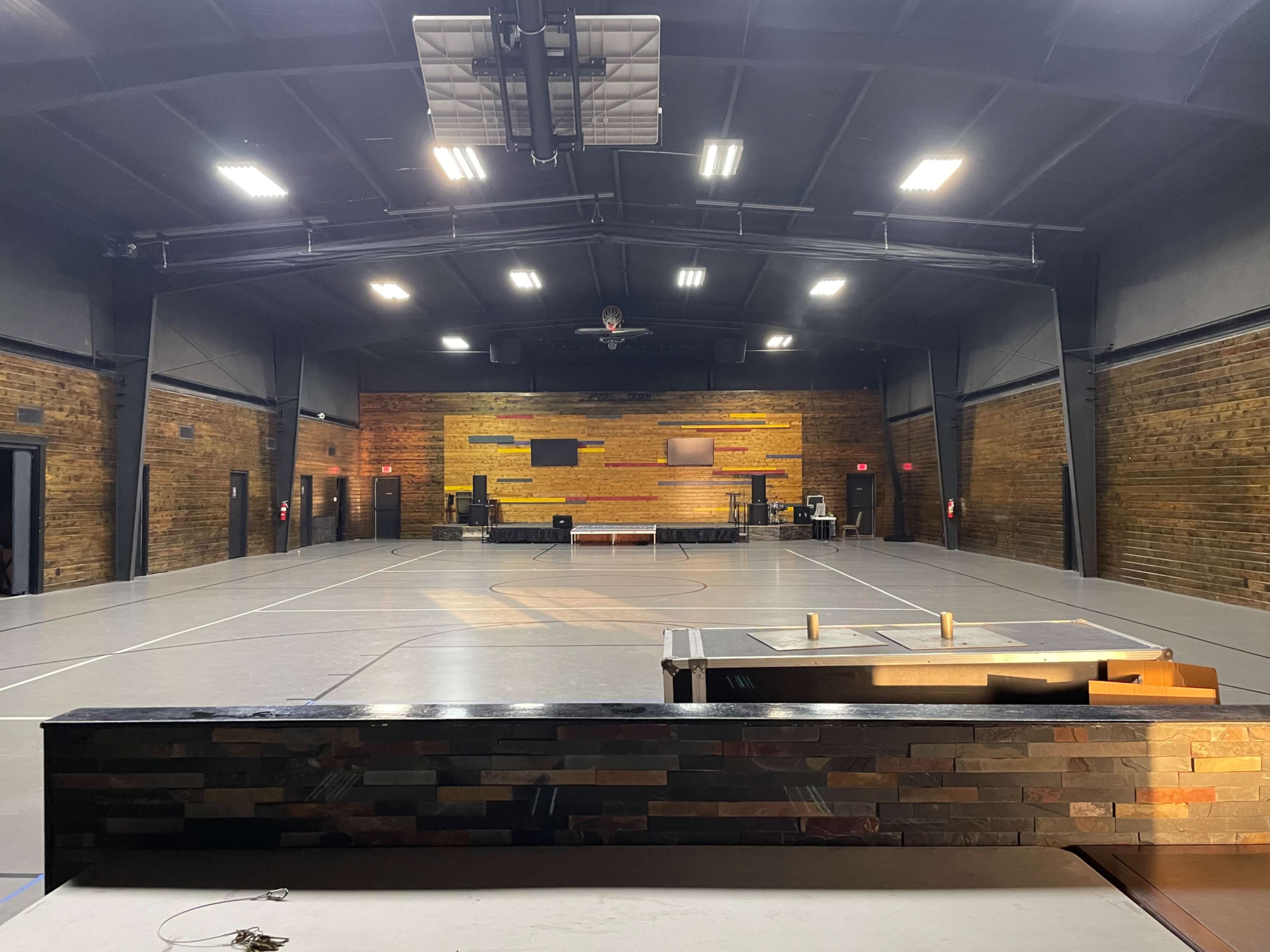 The image shows an empty indoor gymnasium with wooden walls, a sports court, and various equipment at the far end.