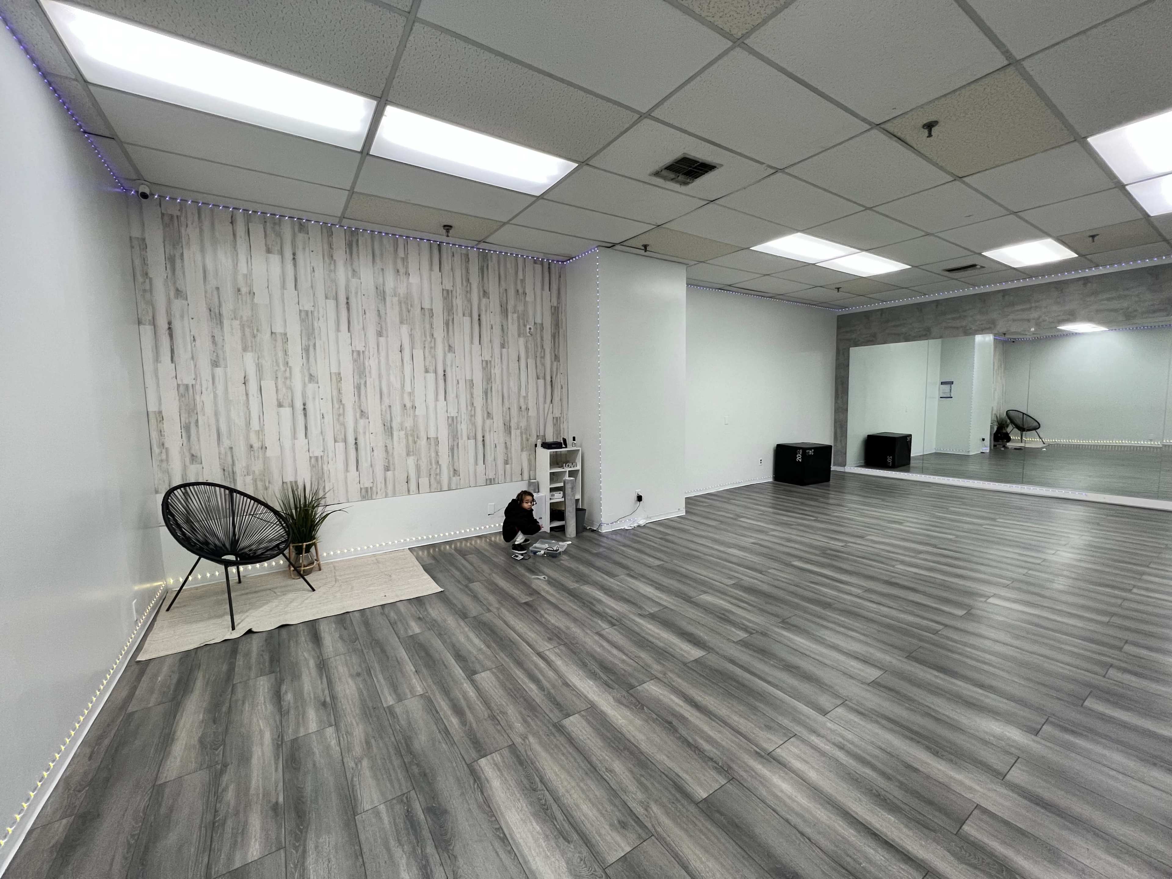 The image depicts a spacious, empty studio with light gray wood flooring, a decorative wall featuring wood paneling, and a small black chair in a corner.