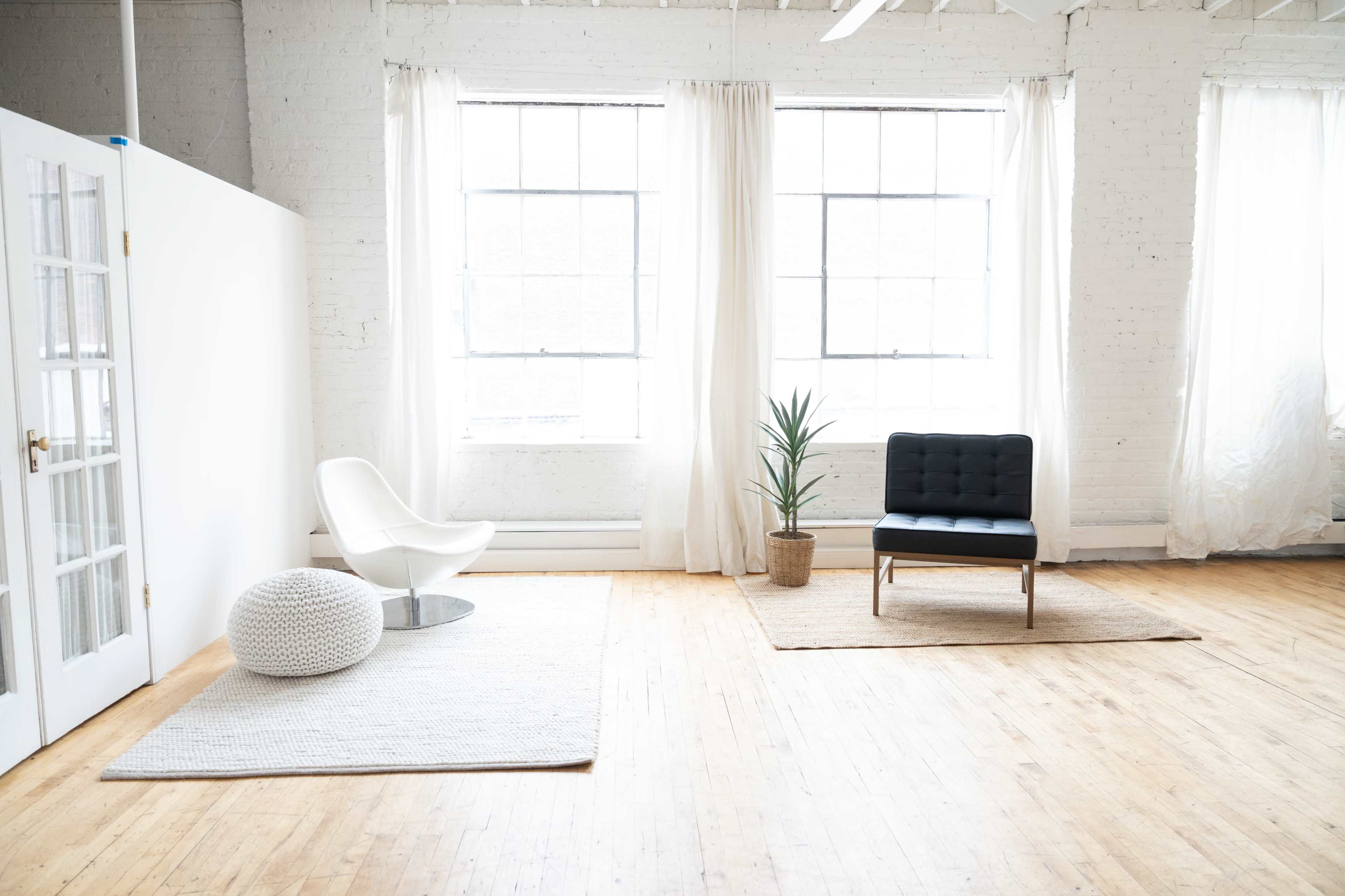 The image shows a spacious room with large windows, featuring a white chair, a black couch, a potted plant, and rugs on a wooden floor.