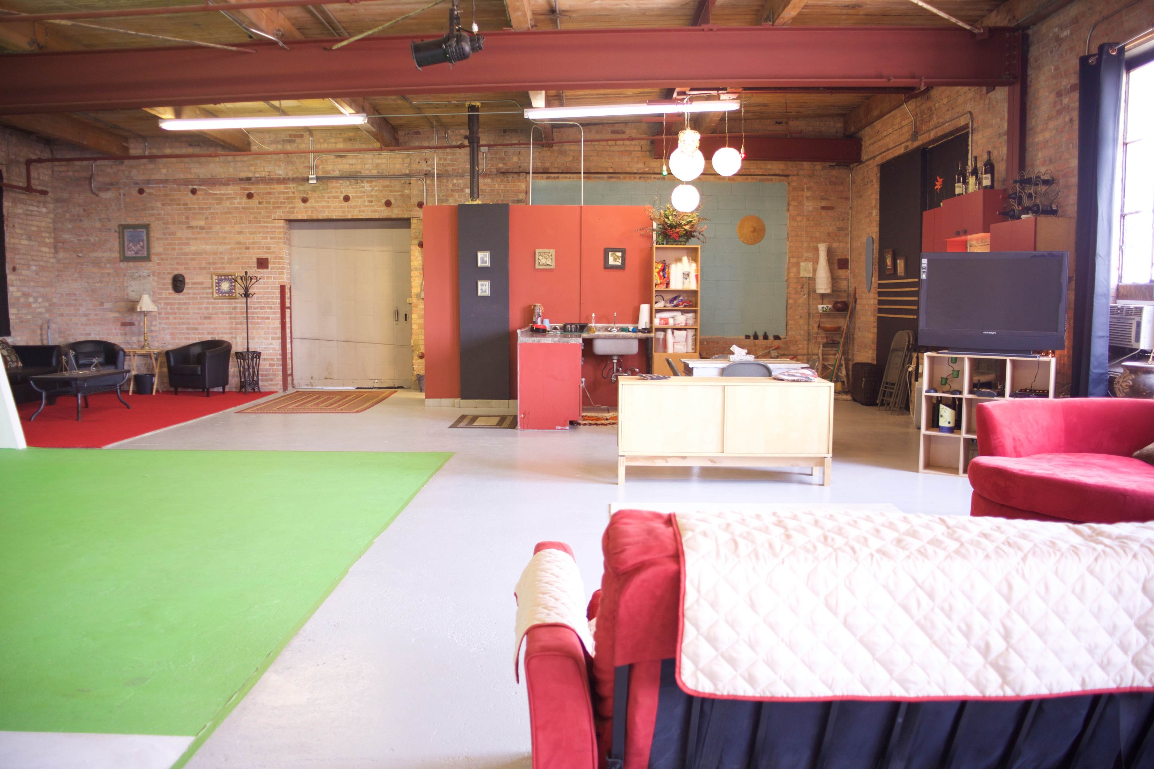 A spacious loft interior with exposed brick walls, a green area rug, a red seating area, and a kitchen setup in the background.