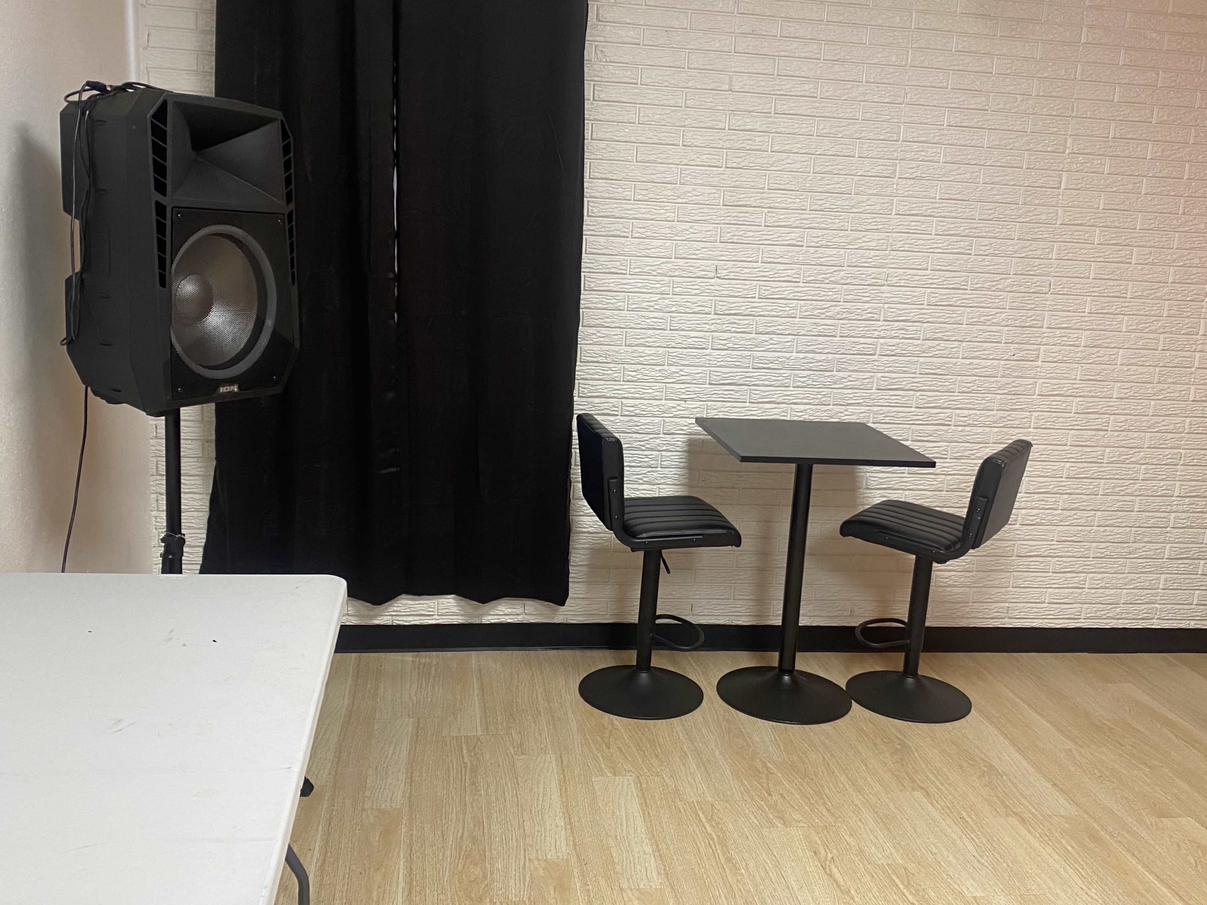 A small room with a speaker on the left, a black table, and two chairs against a white brick wall.