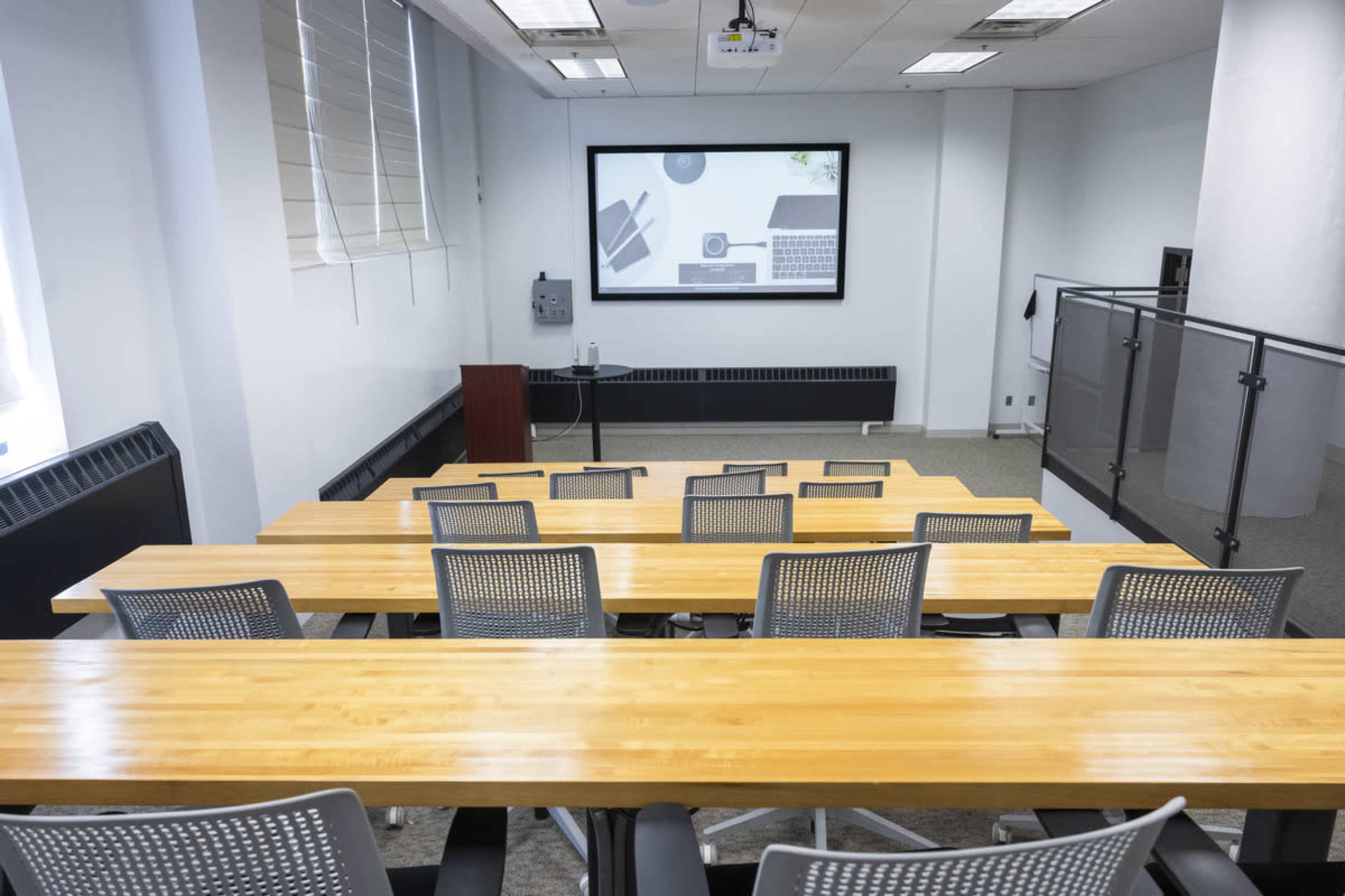 A conference room features rows of wooden tables and mesh-backed chairs, with a large screen displaying graphics in the background.