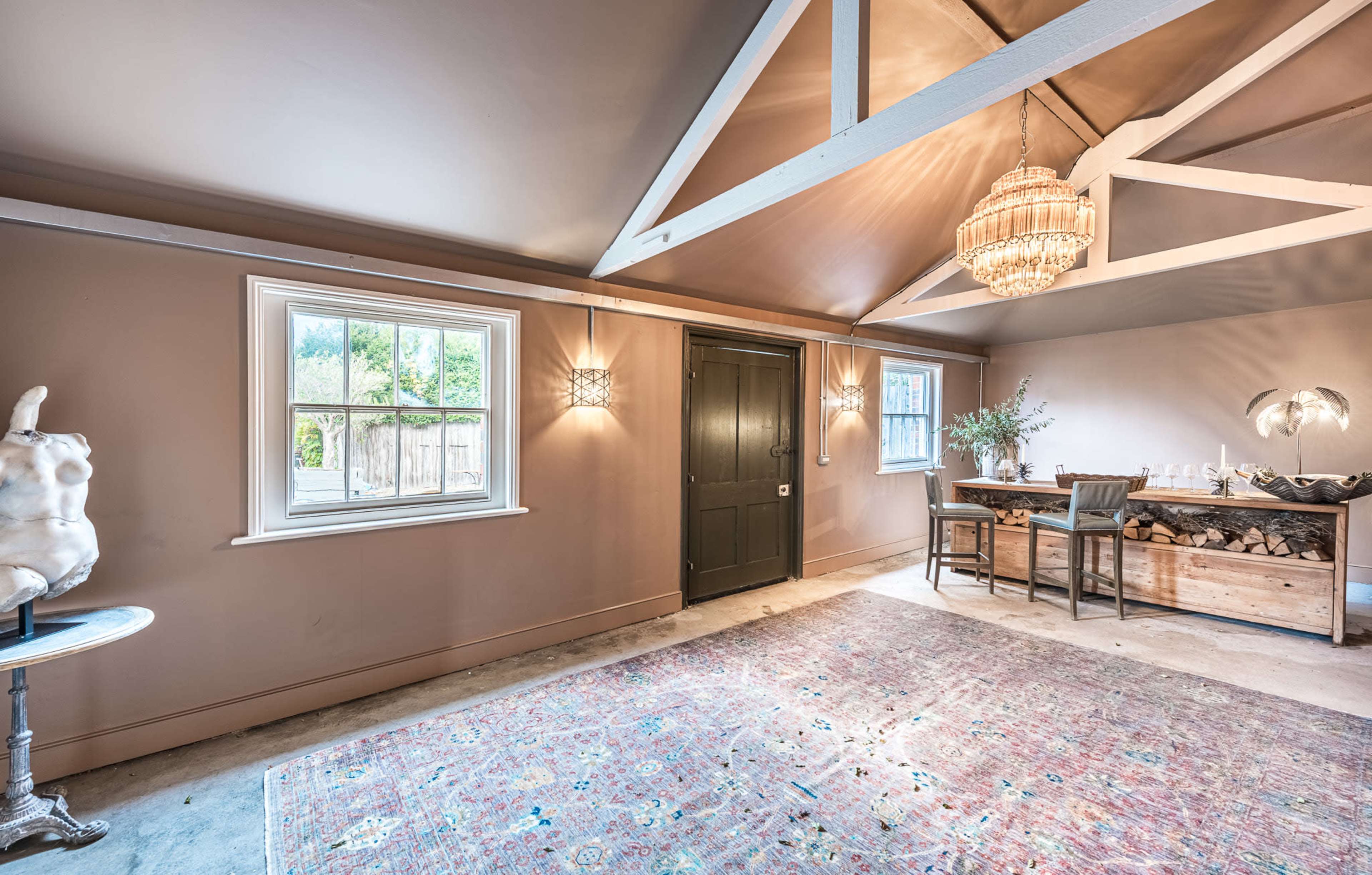 The interior of a room features a chandelier, a table with decorative items, and a large window, with light-colored walls and exposed beams.