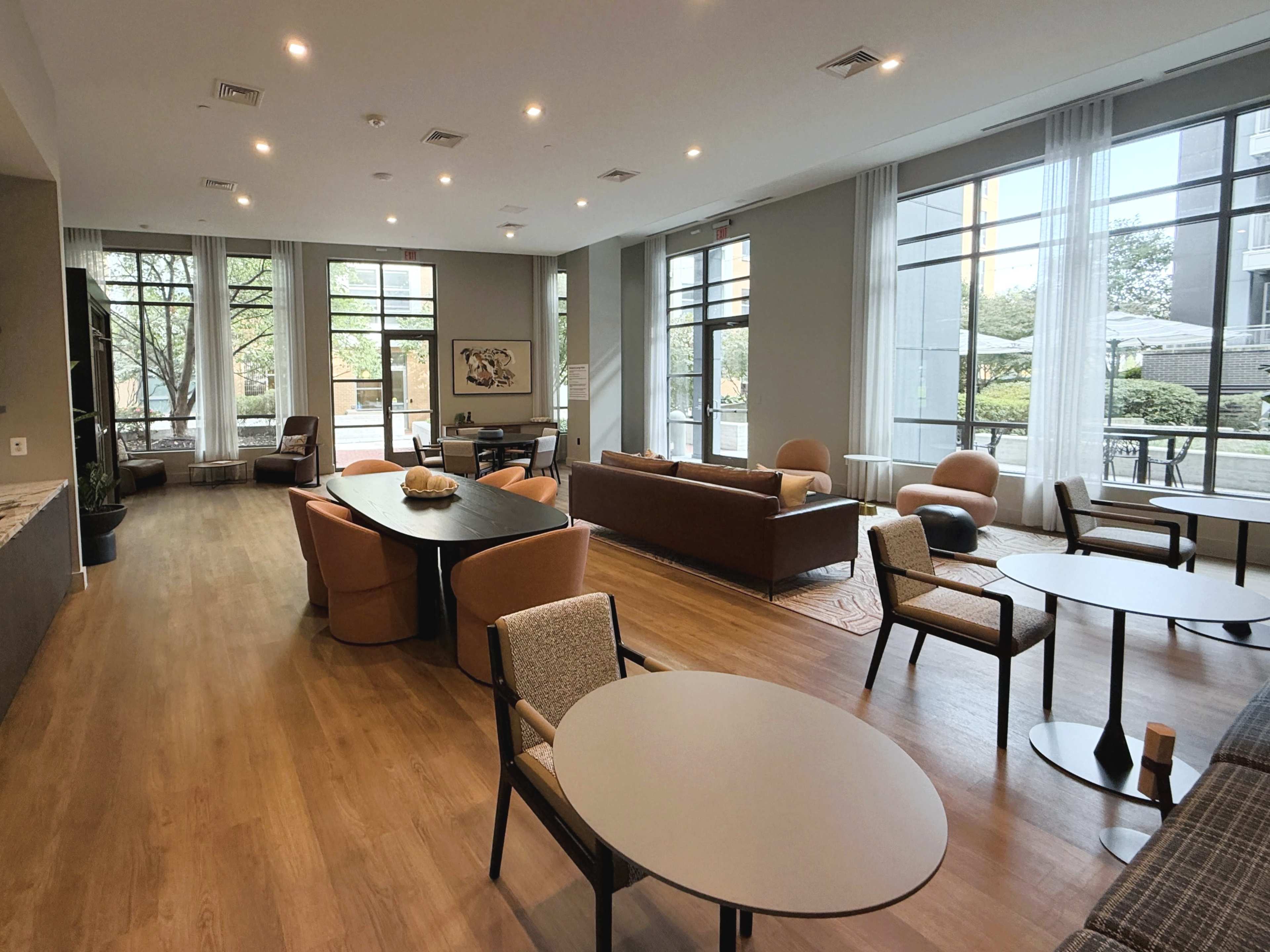 The image shows a modern lounge area with seating arrangements, tables, and large windows allowing natural light to enter.