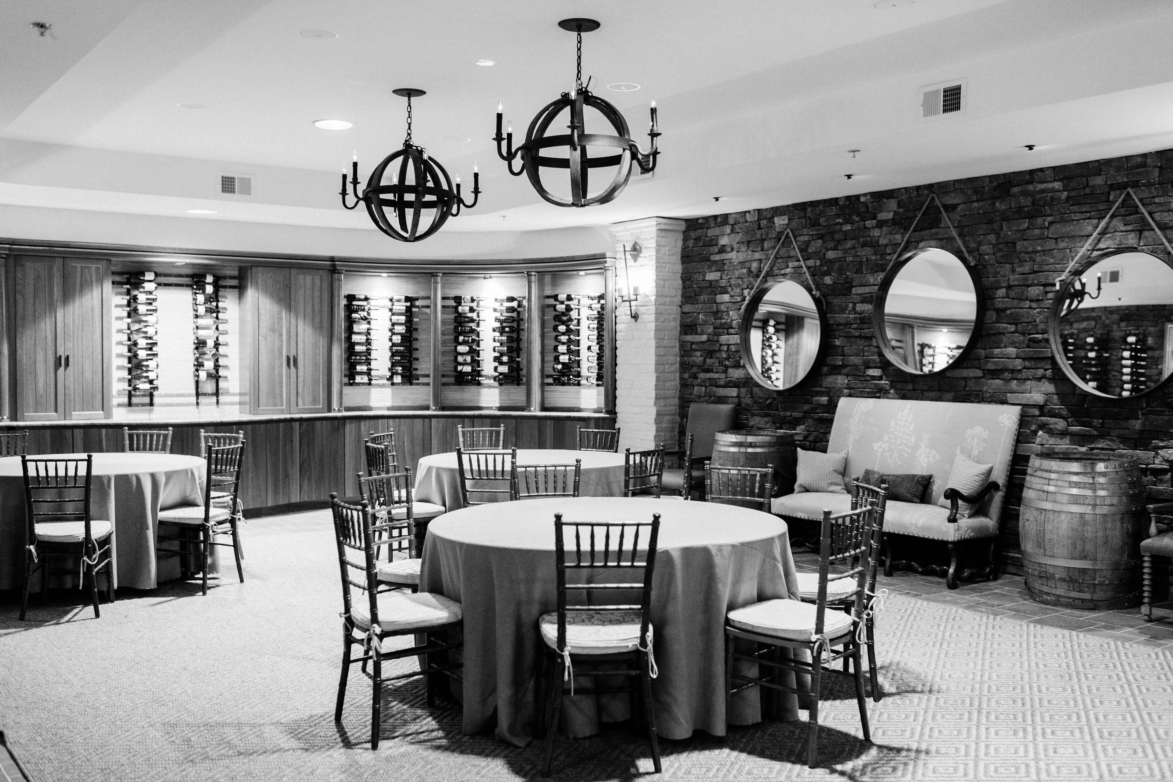 The room features several round tables surrounded by chairs, with a wine display mounted on the wall and mirrors reflecting the space.