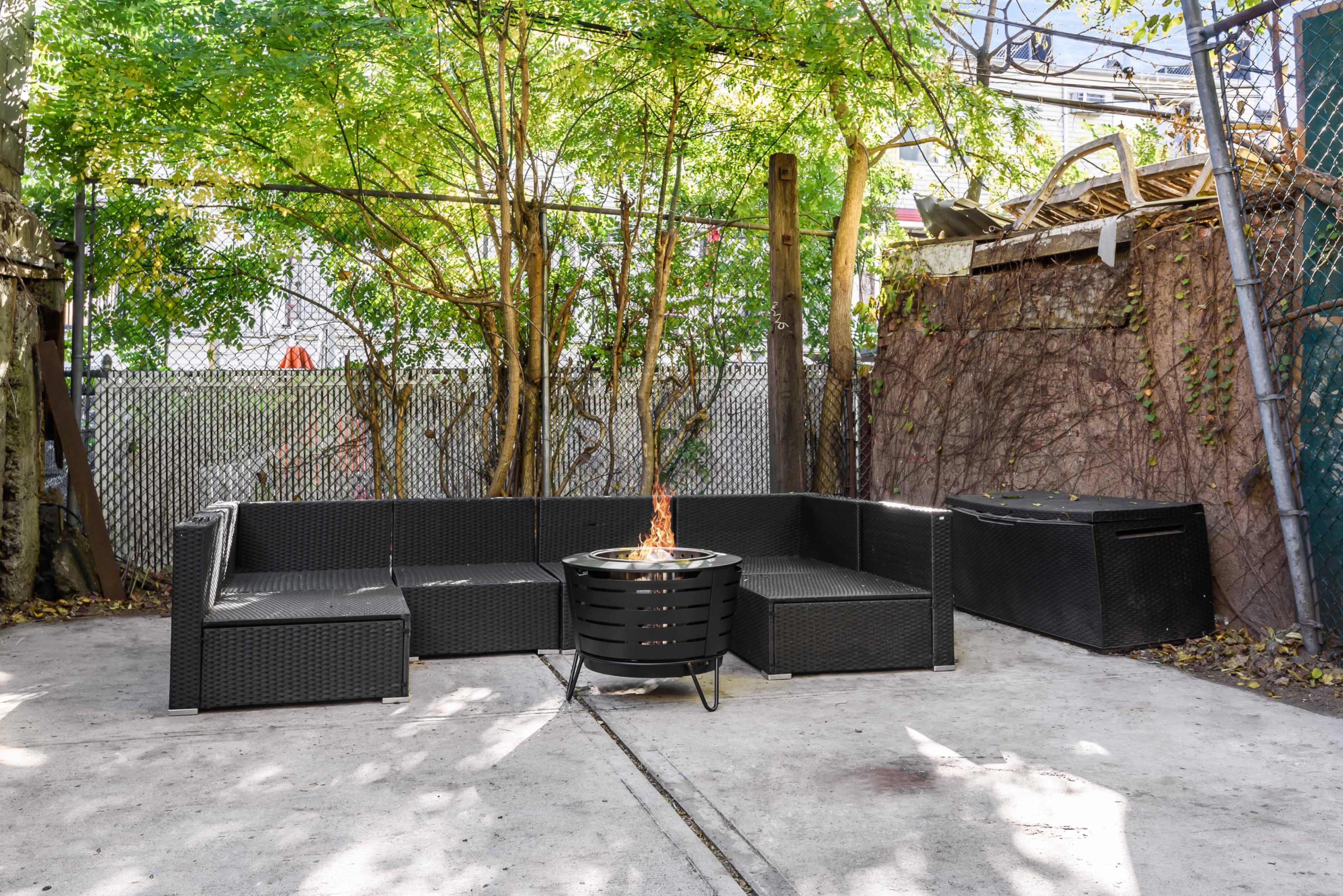 A patio area features a black wicker seating arrangement surrounding a circular fire pit, set against a backdrop of greenery and a fenced wall.