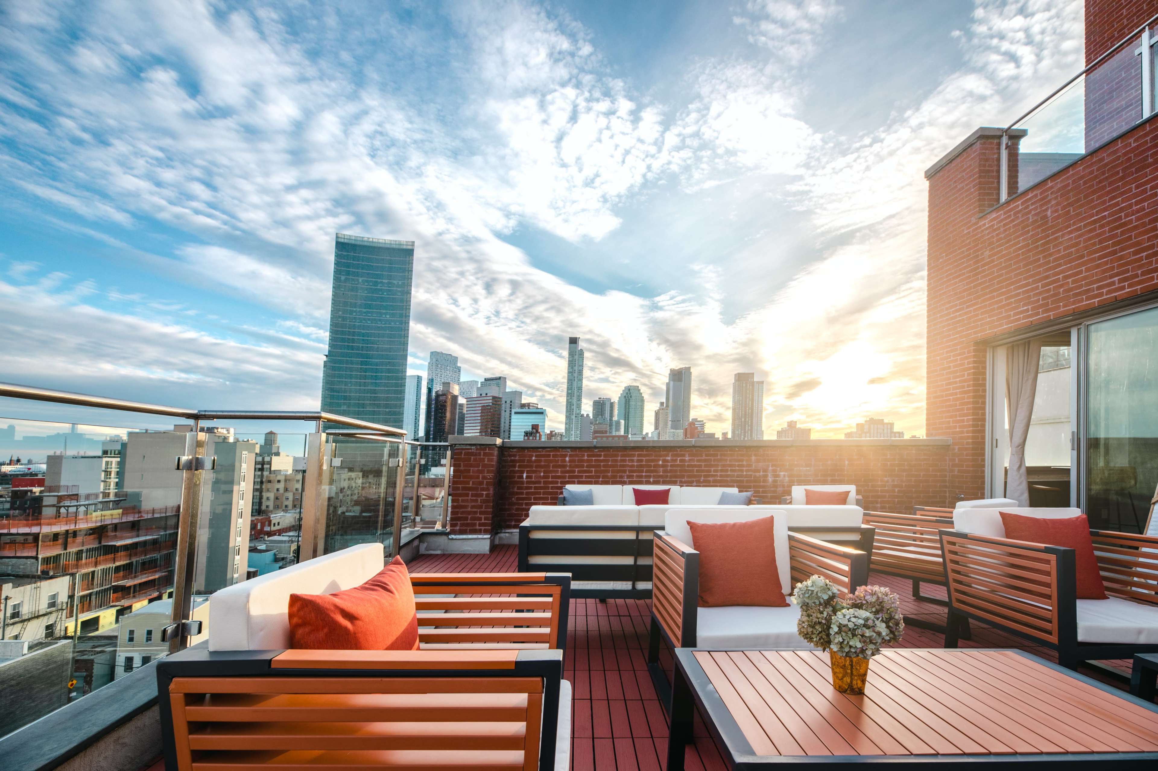 A rooftop terrace features modern seating arrangements with orange cushions and a view of a city skyline at sunset.