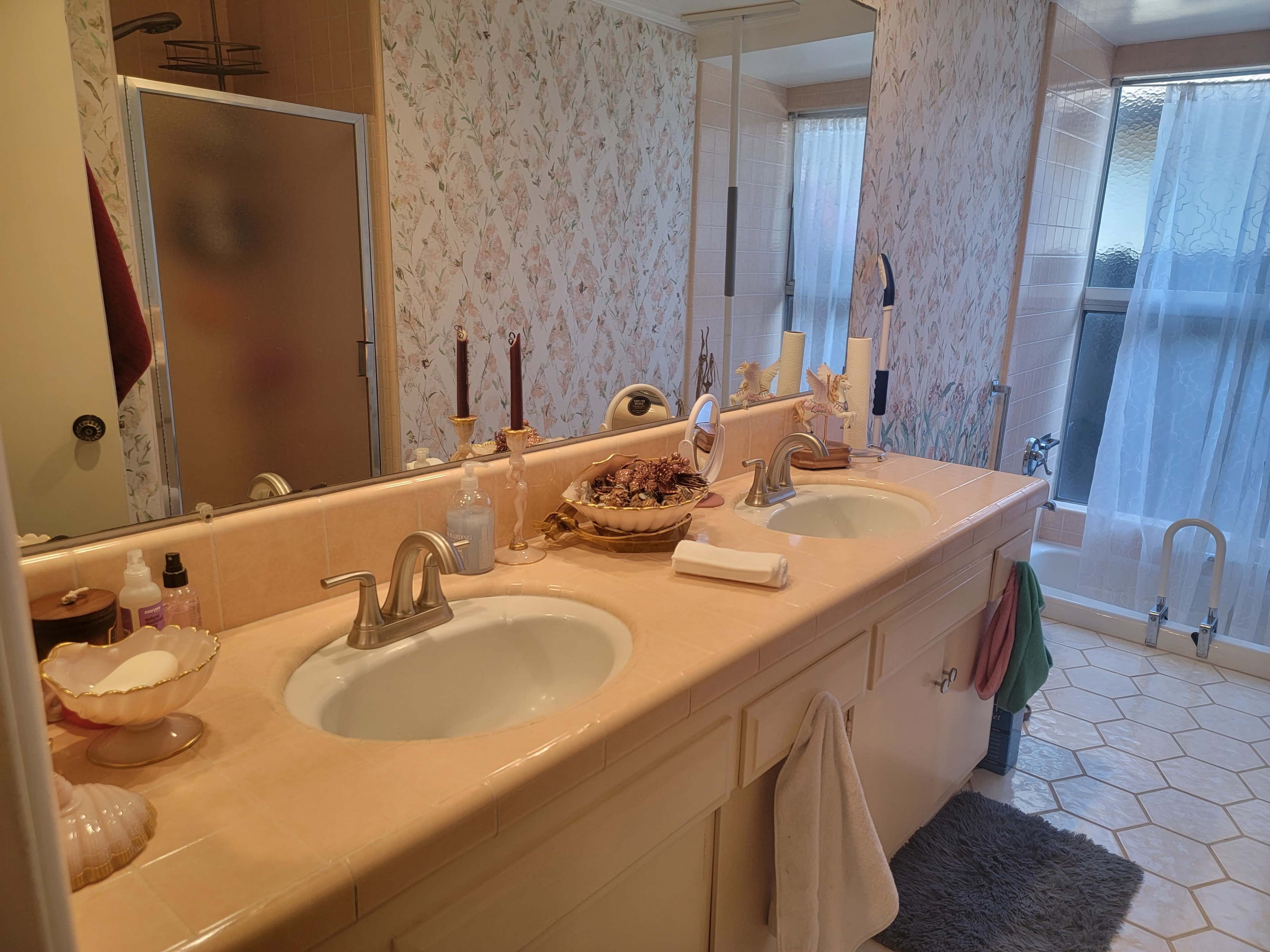 A bathroom with a double sink vanity, decorative wallpaper, and a bathtub visible through a glass door.