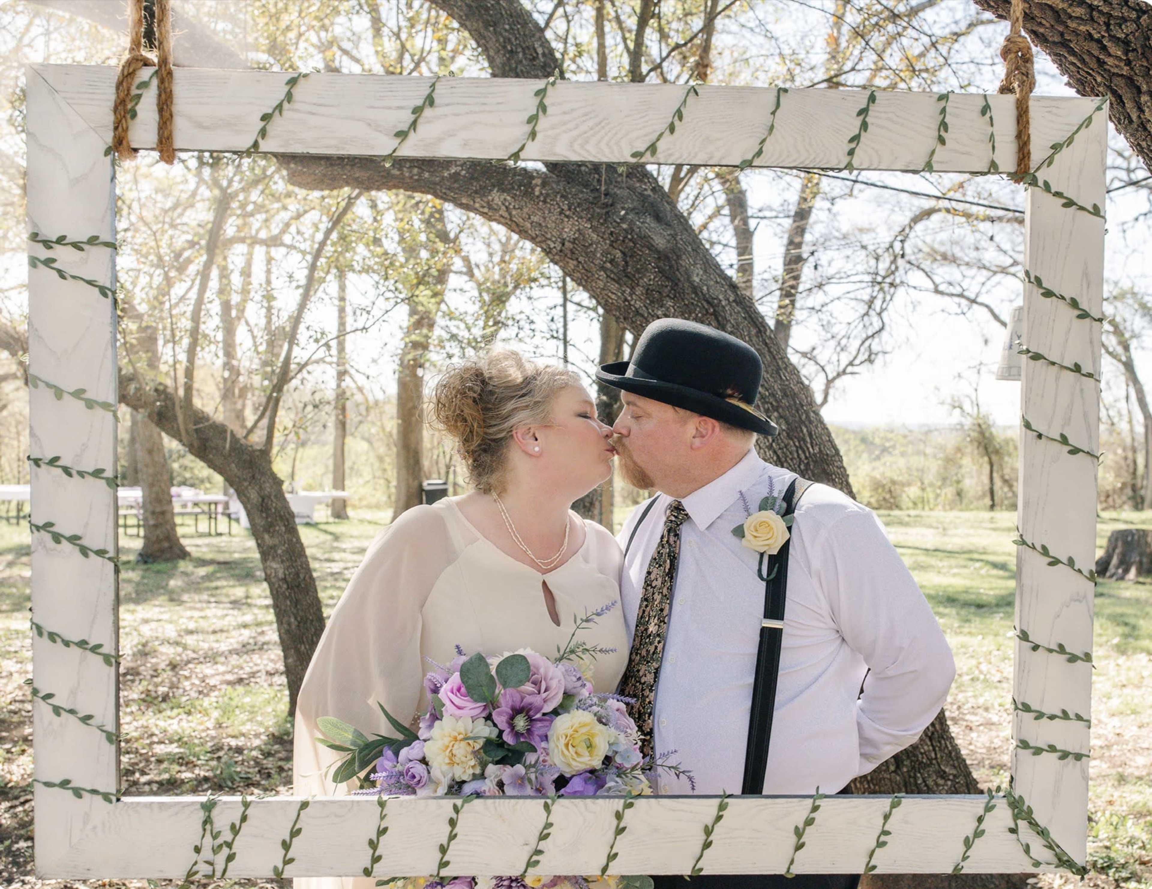 A couple dressed in formal wedding attire kisses inside a large decorative frame hanging from a tree in a natural outdoor setting.