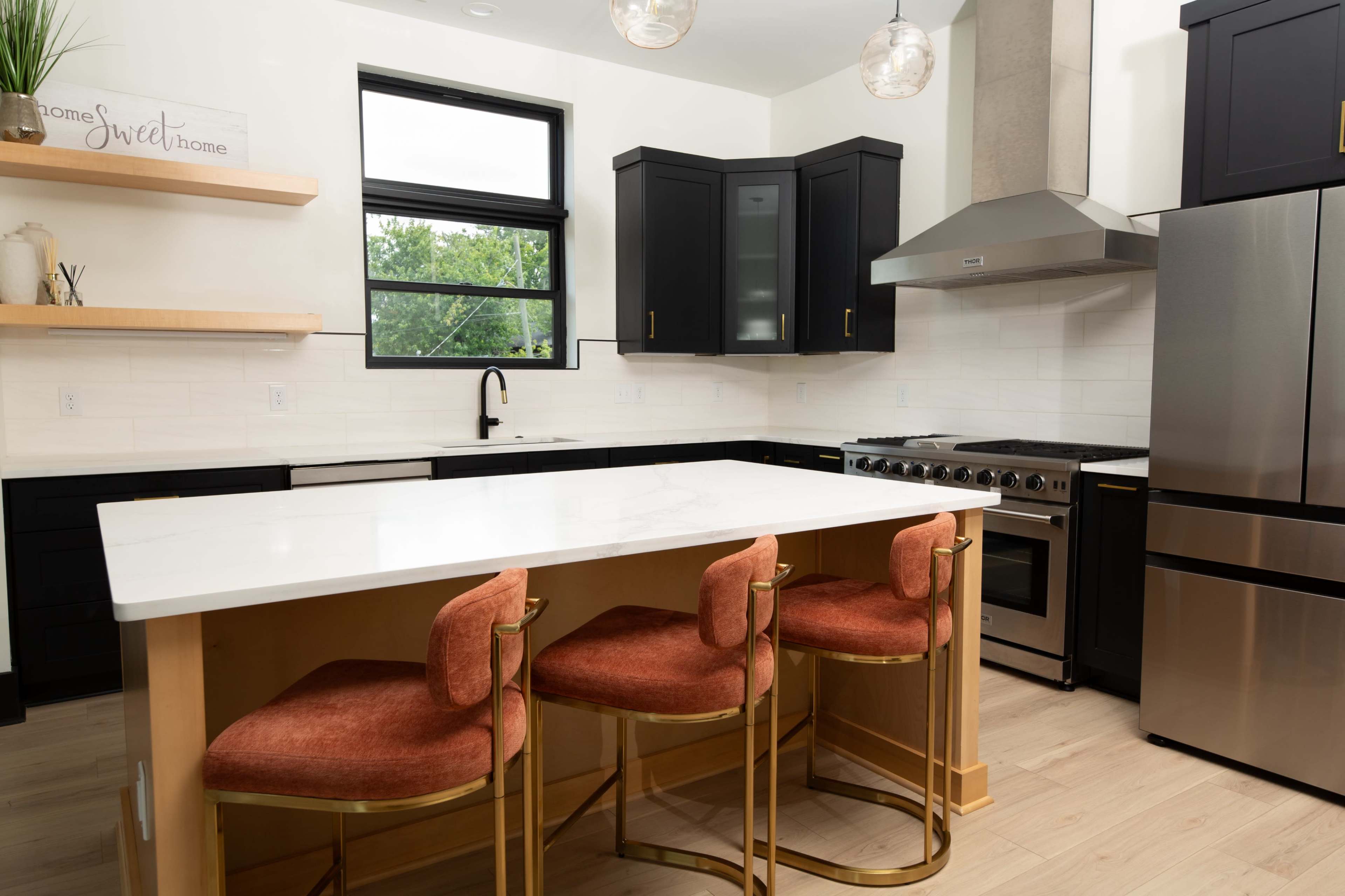 The image shows a modern kitchen with black cabinetry, a stainless steel stove, and a marble countertop island featuring three gold-accented bar stools.