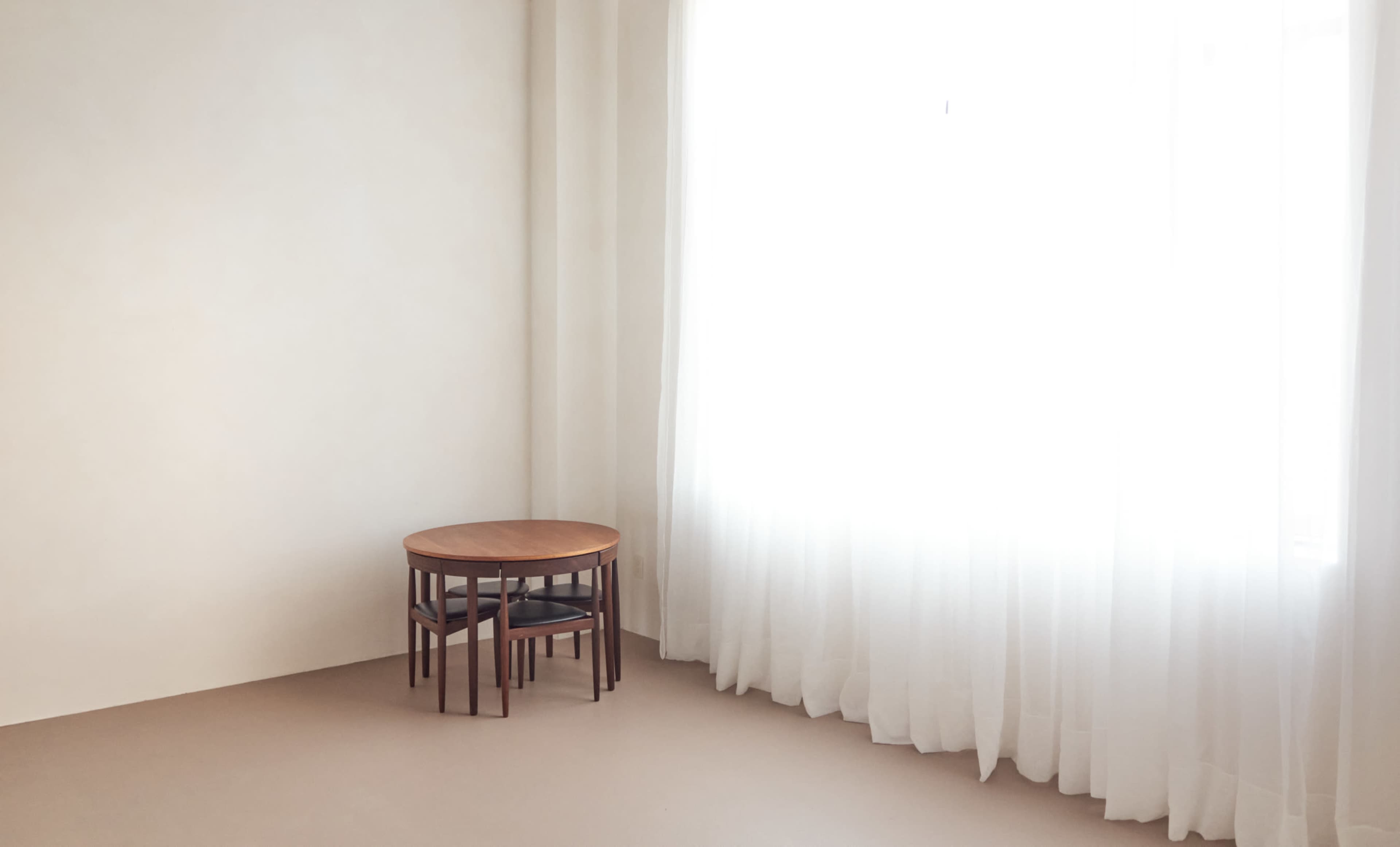 A small wooden table with chairs stacked underneath is positioned next to a large window covered with sheer curtains.
