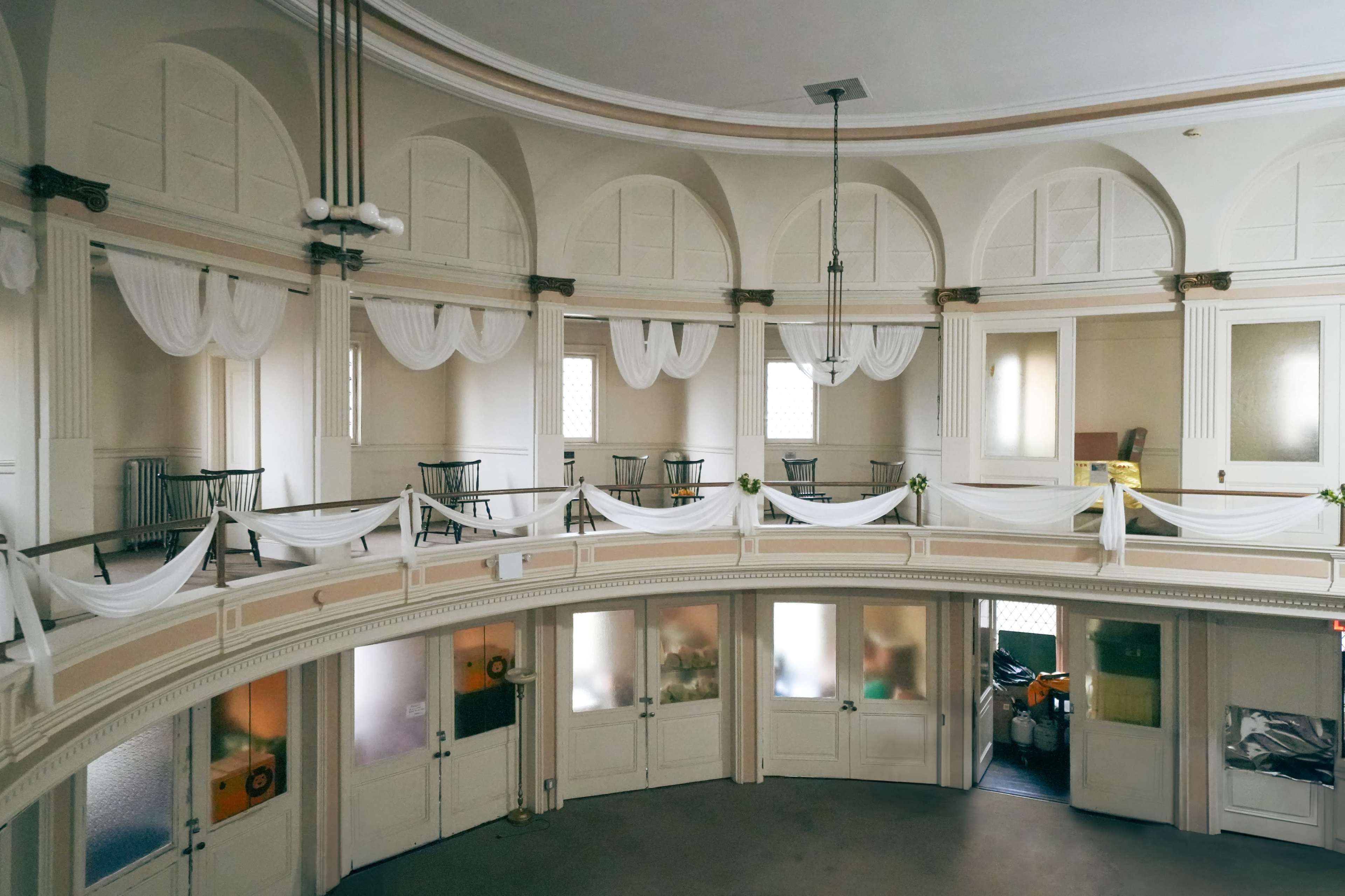 An interior space with curved architecture, featuring a series of arched windows and white draped fabric, along with tables arranged on a balcony level.