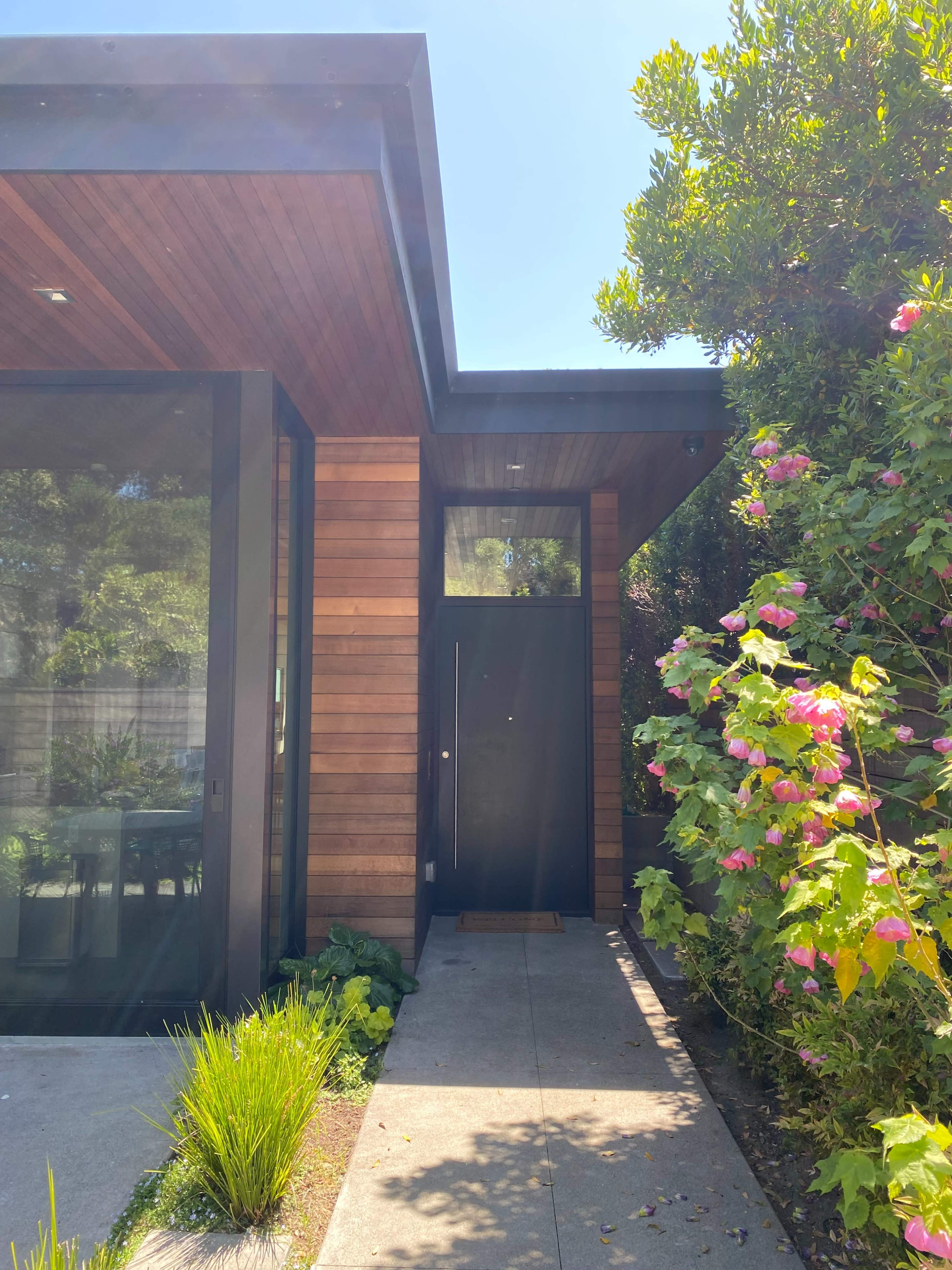 A modern house entrance features a wooden door surrounded by greenery and colorful flowers.