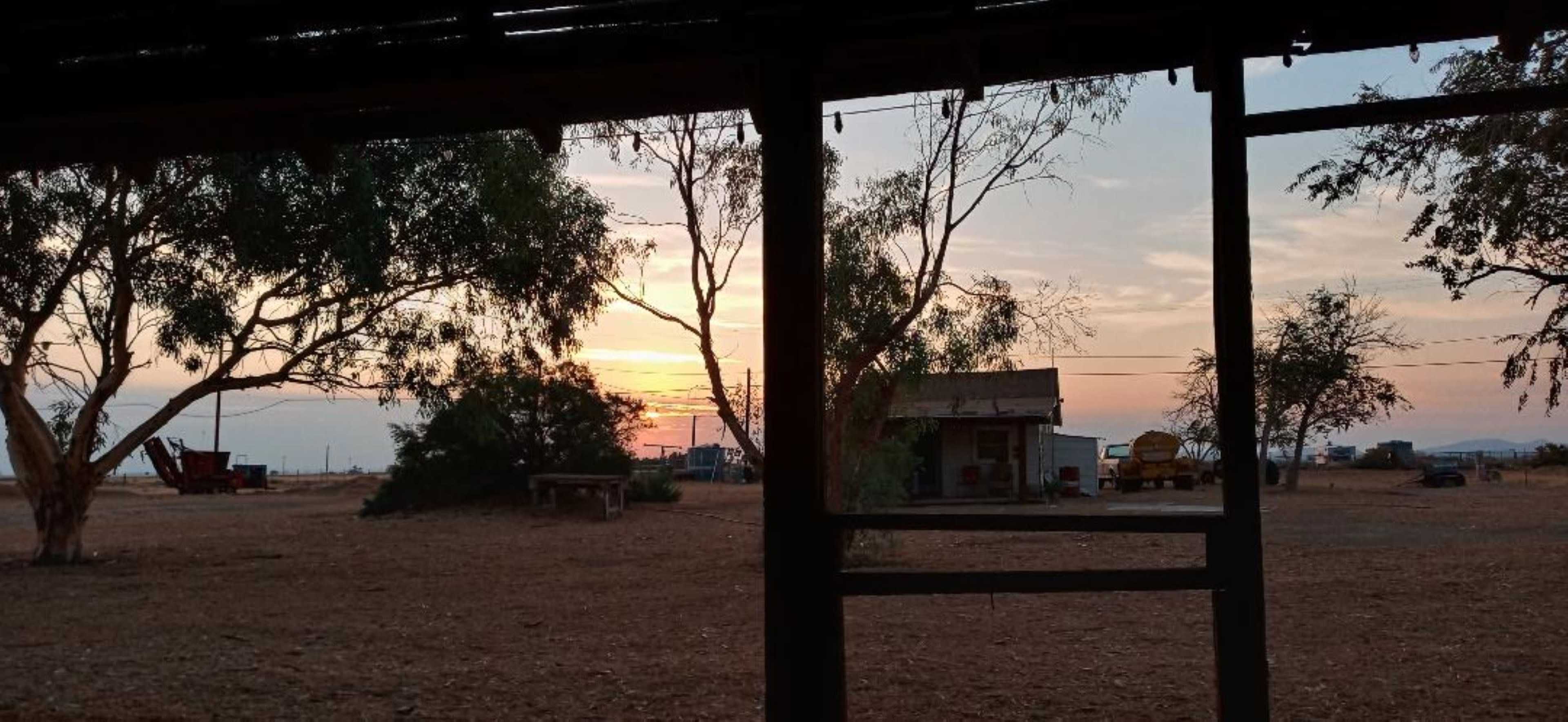 A sunset viewed through a rustic structure, with silhouettes of trees and machinery in the background.