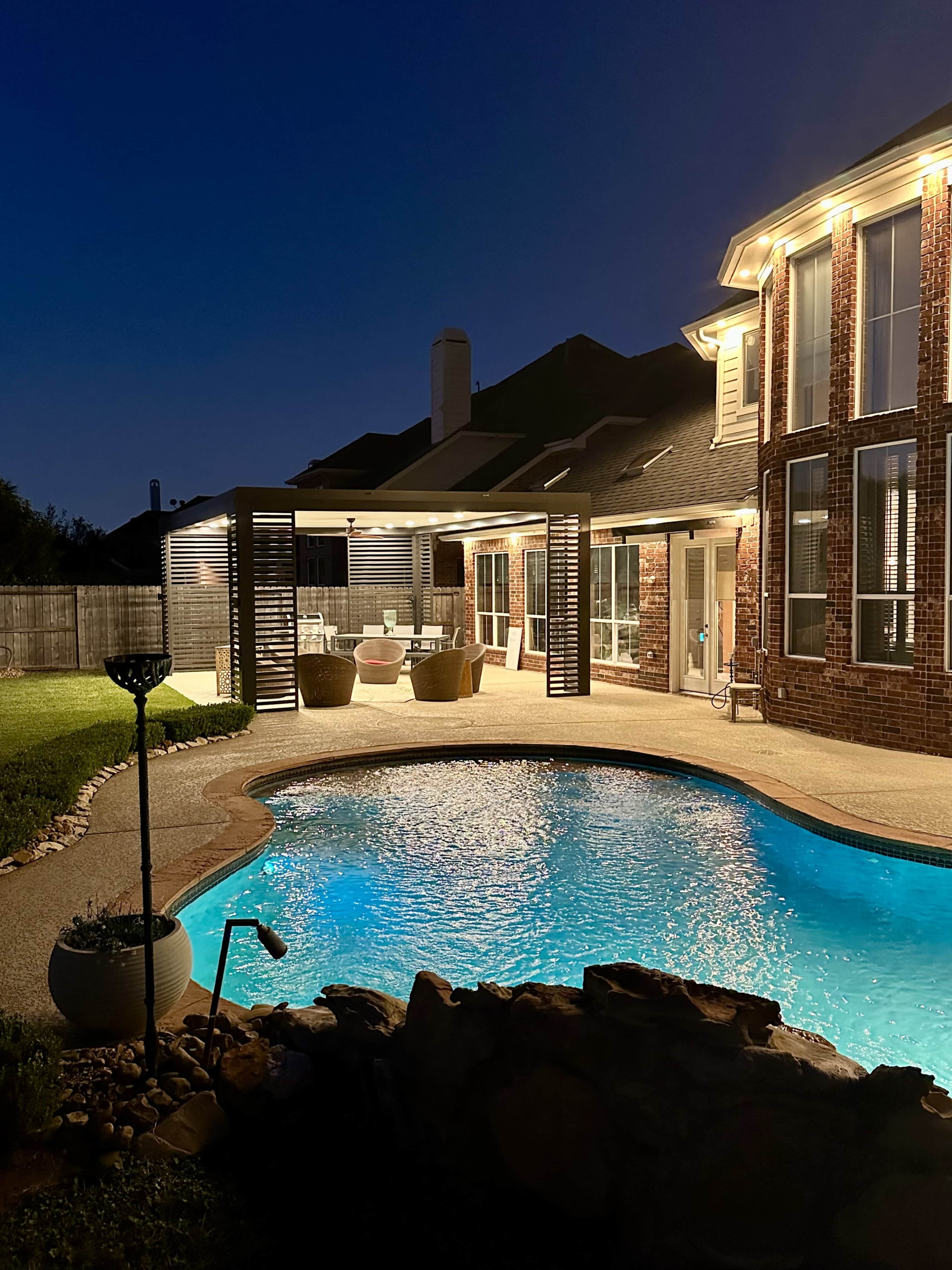 The image shows a well-lit backyard with a swimming pool, a covered seating area, and a brick house at night.