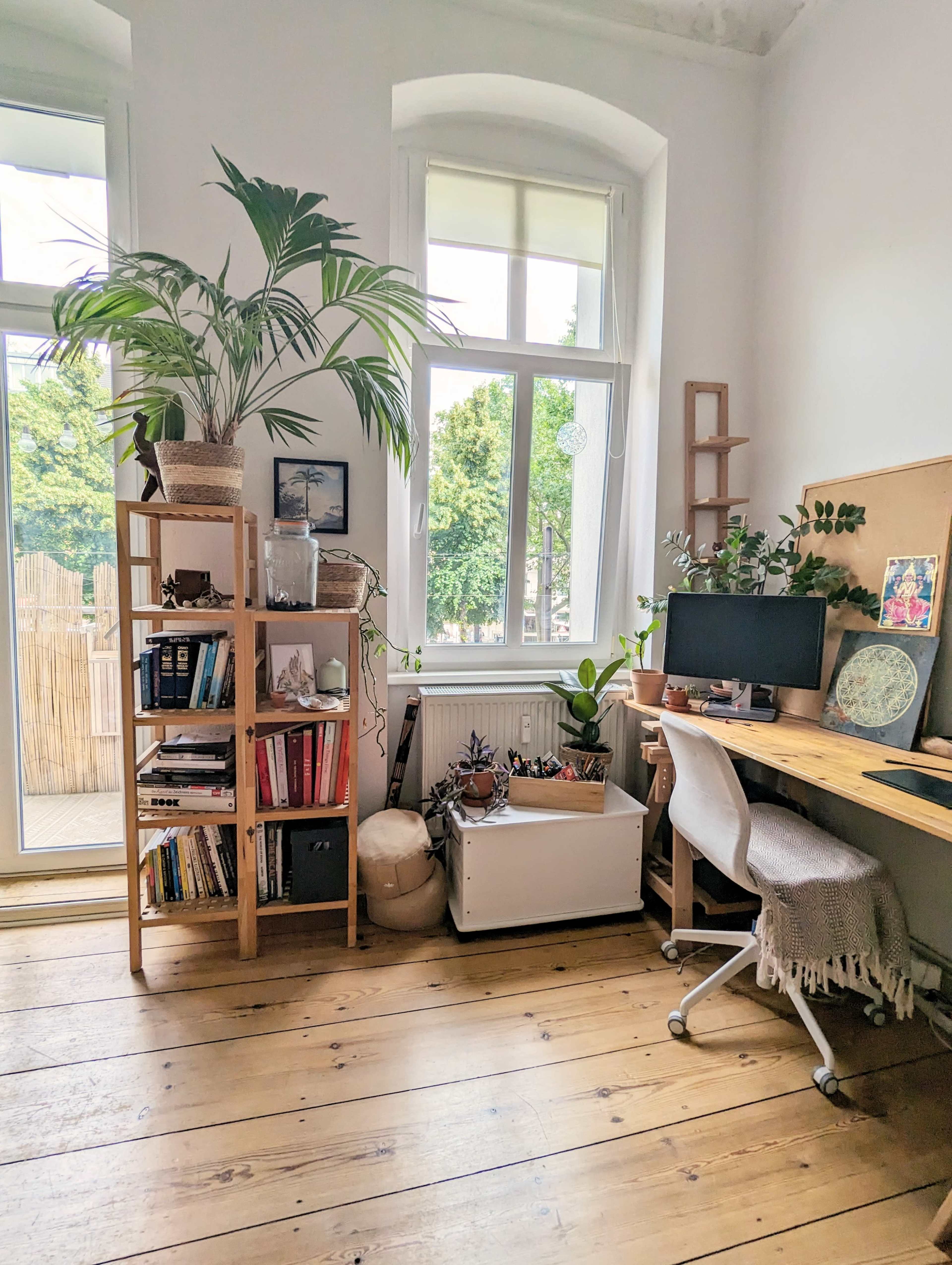 A cozy home office with a wooden desk, a computer, a bookshelf filled with books, and indoor plants near a large window.