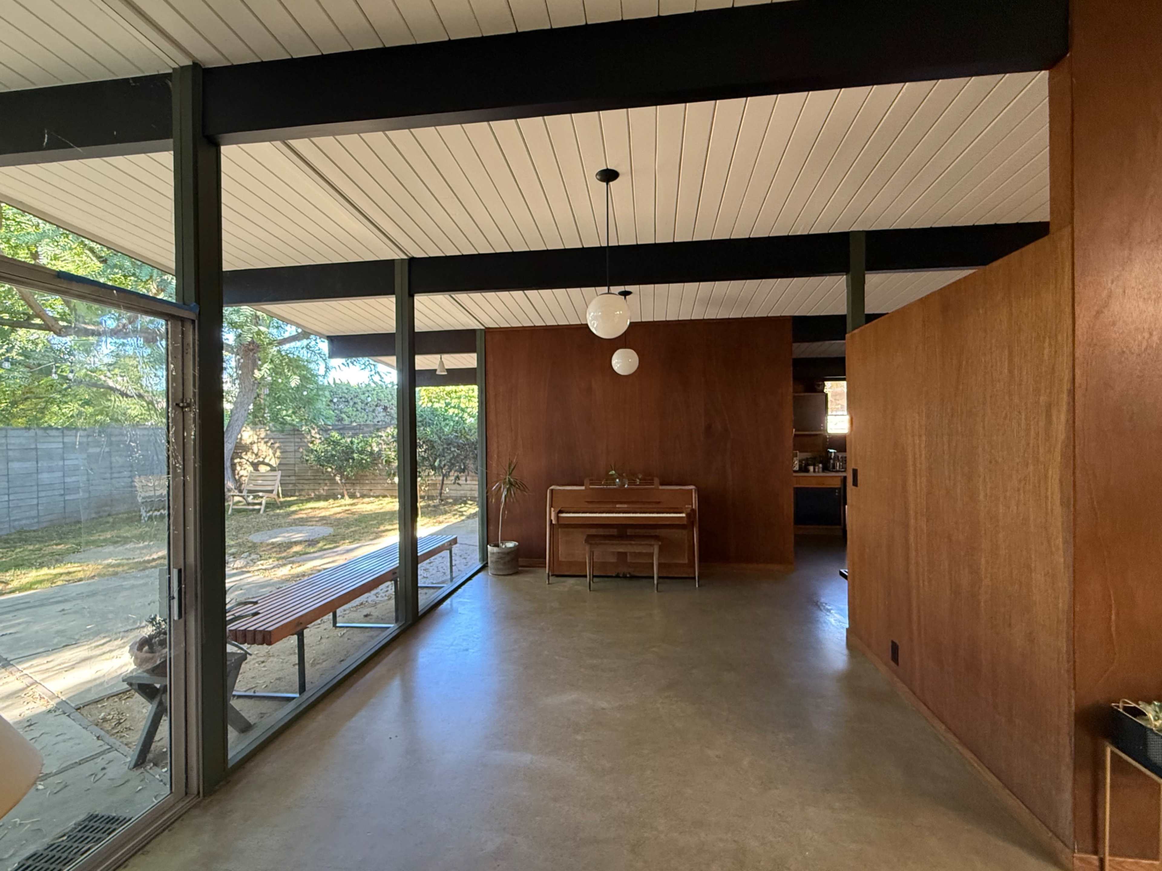 The image shows an interior space of a modern house featuring wooden walls, large windows, and a piano against one wall.