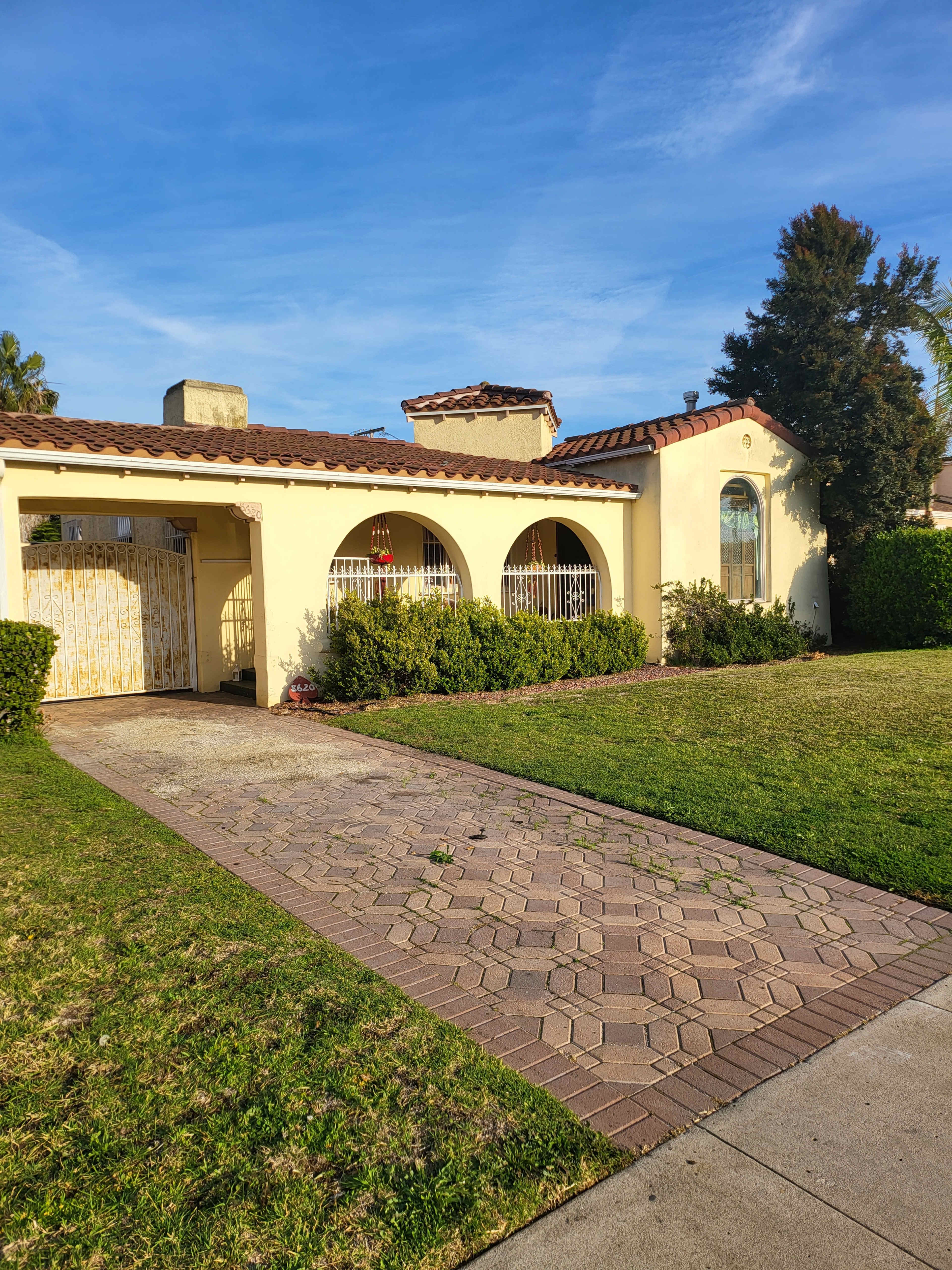 The image shows a single-story, yellow stucco house with a red tile roof and a brick pathway leading to the entrance.