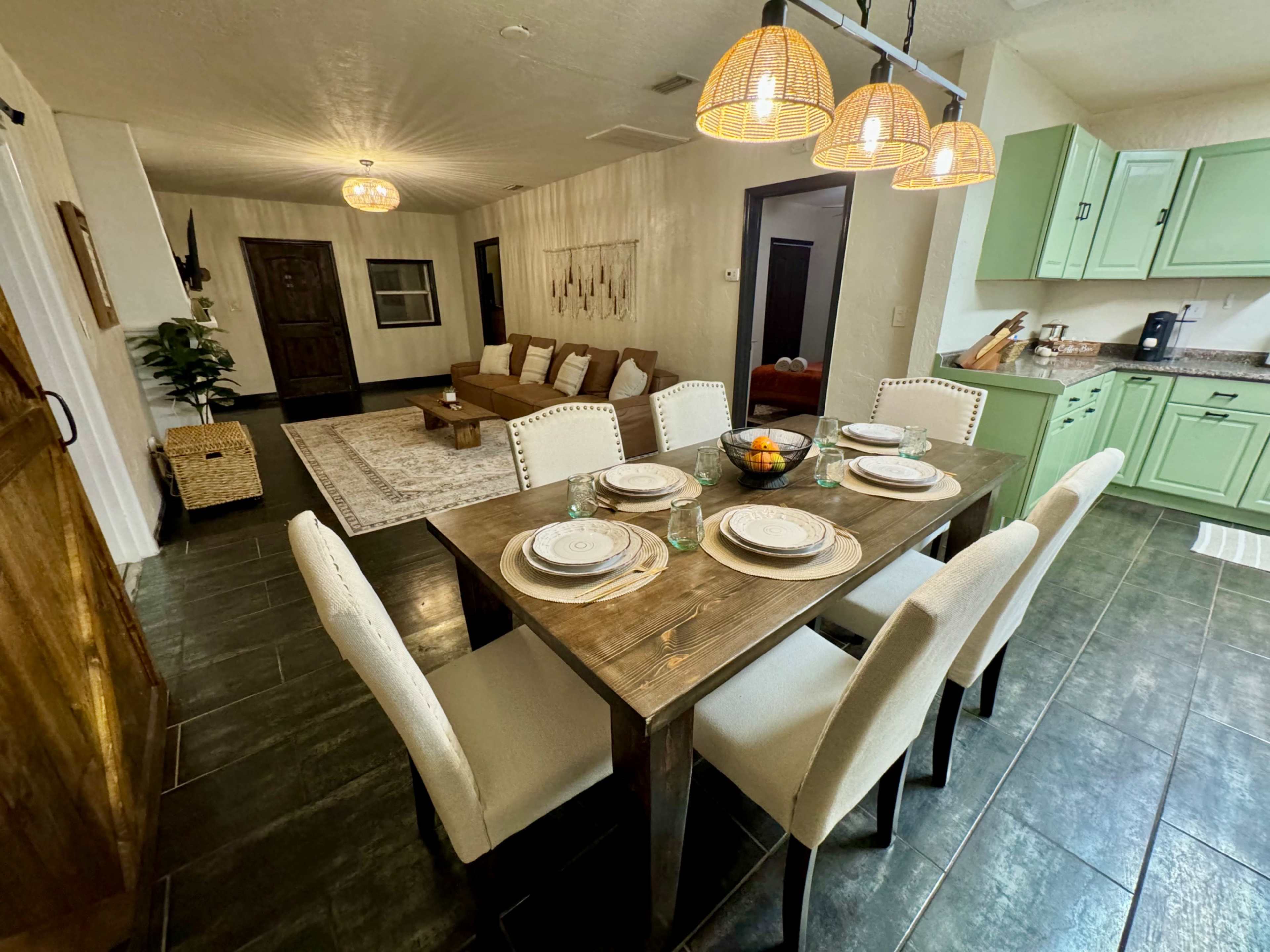 A dining area features a wooden table set for six, with beige chairs, surrounded by a light-filled open space that includes a living area and a kitchen.