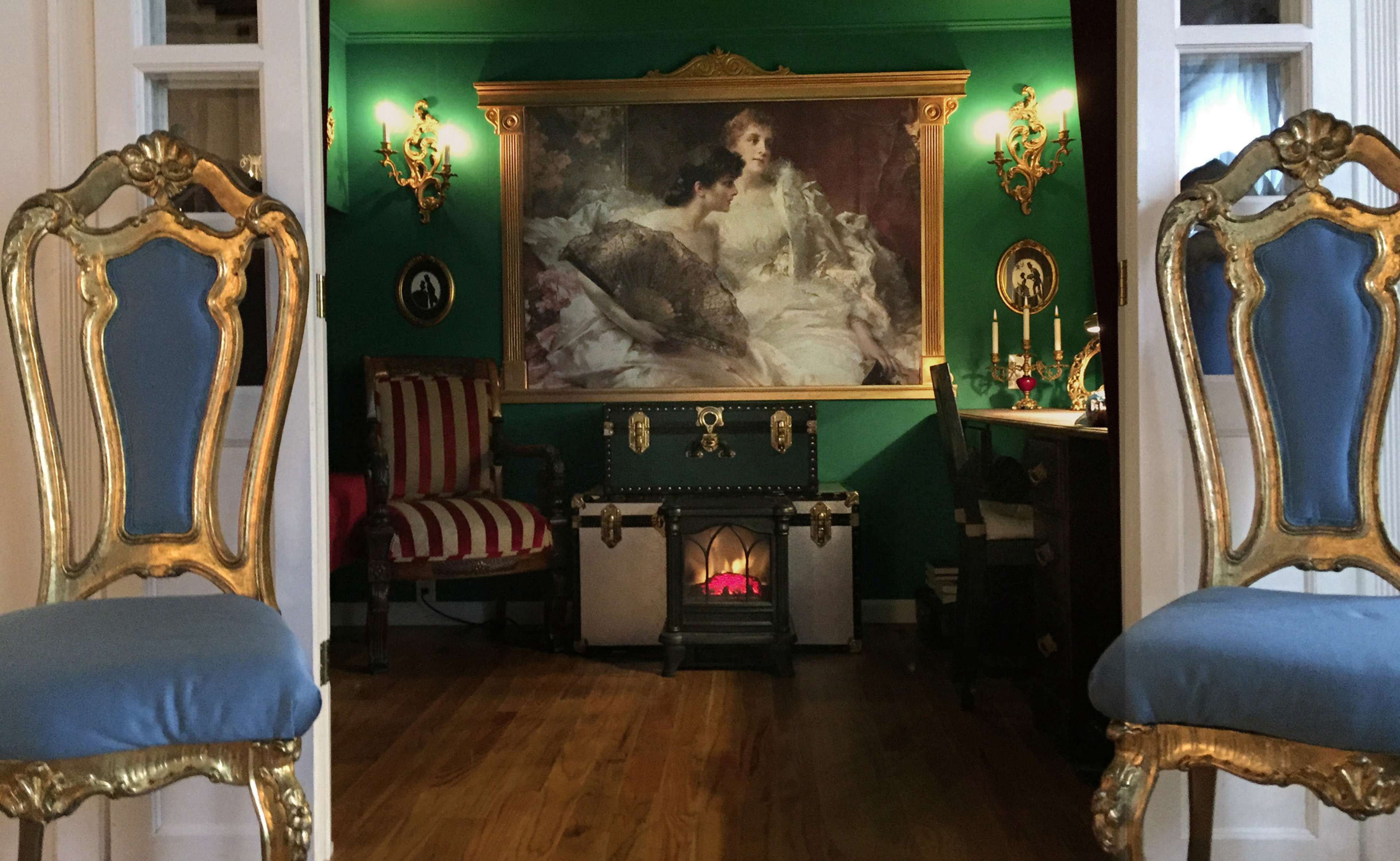 The image shows a room with ornate golden chairs facing a large painting on a green wall, flanked by decorative sconces and a fireplace.